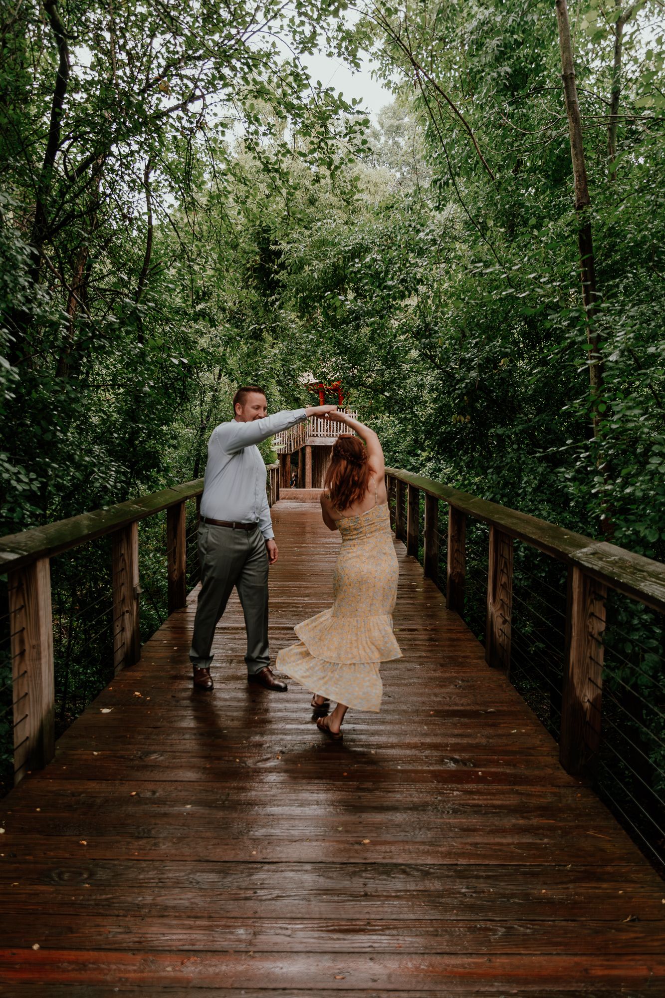 A couple dances together on a wooden boardwalk surrounded by lush green forest during their engagement session.