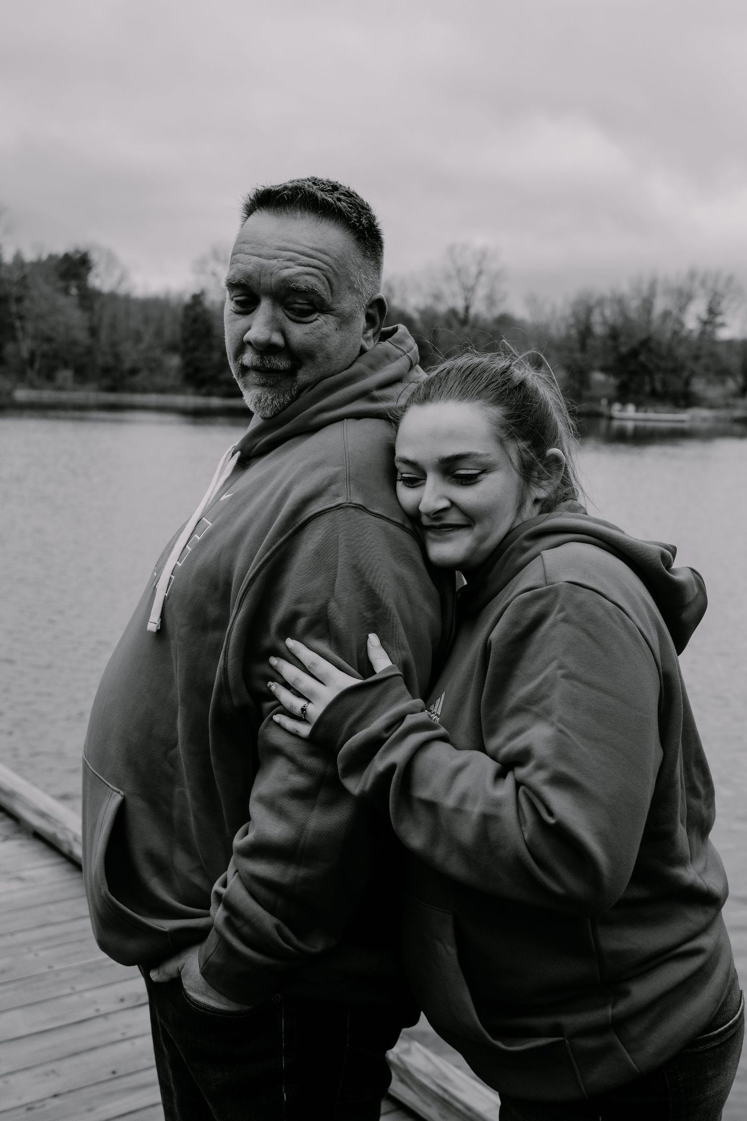 A couple in matching hoodies embrace on a wooden dock by a lake.