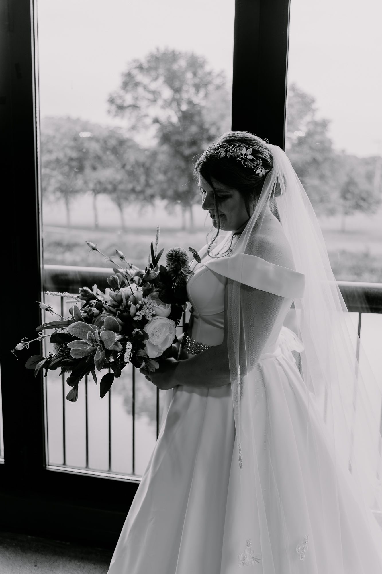 Black and white portrait of bride holding bouquet by window in elegant wedding dress.