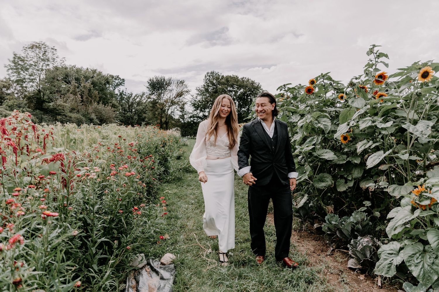Newlyweds walking hand in hand through garden path with blooming flowers.