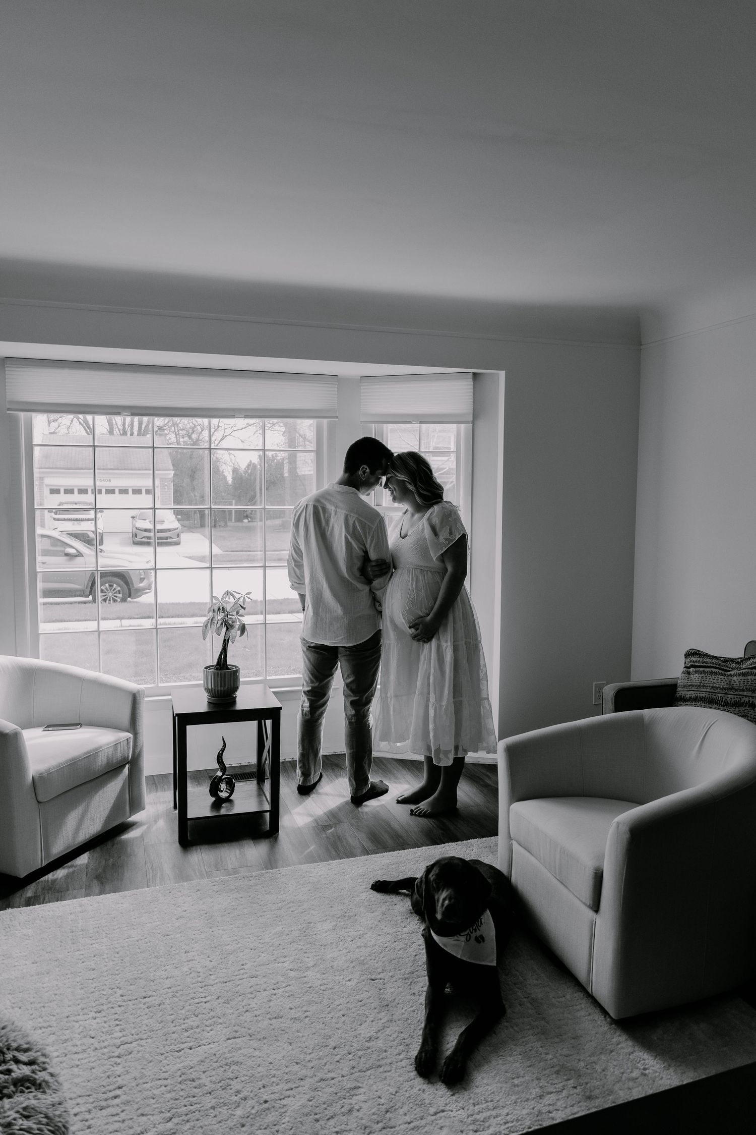 Black and white photo of a couple sharing a tender moment by a large window, with a dog resting on a rug nearby.
