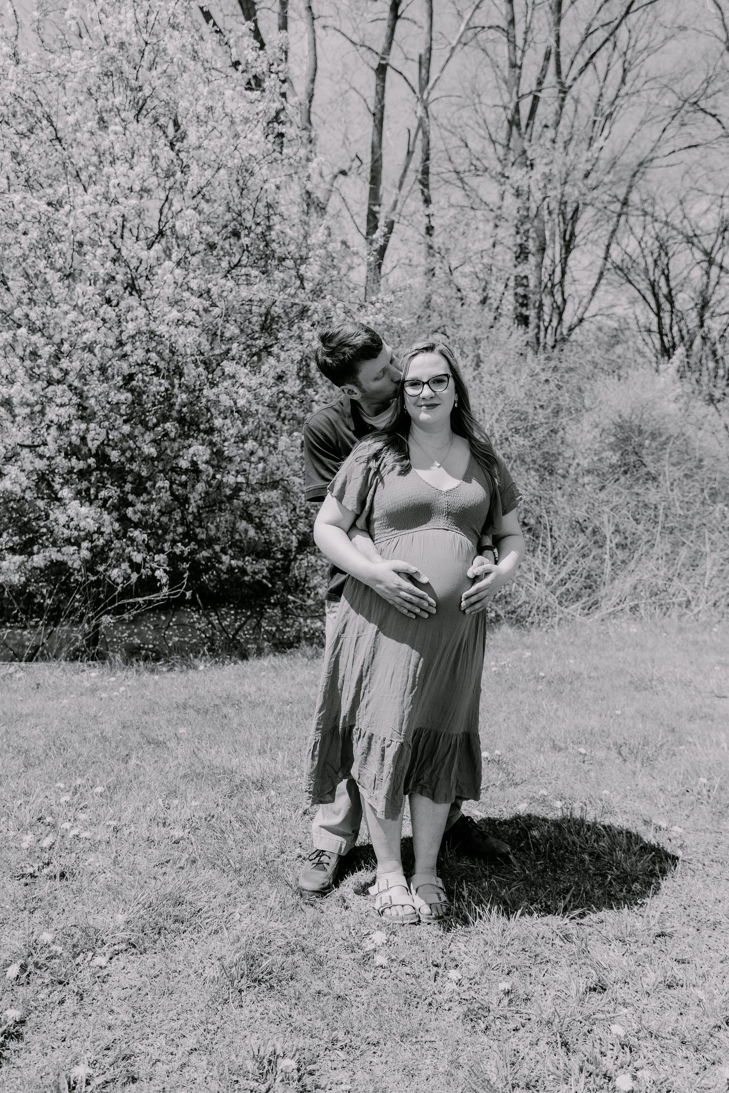 Black and white maternity photo of a couple outdoors, man embracing pregnant woman from behind near blooming trees.