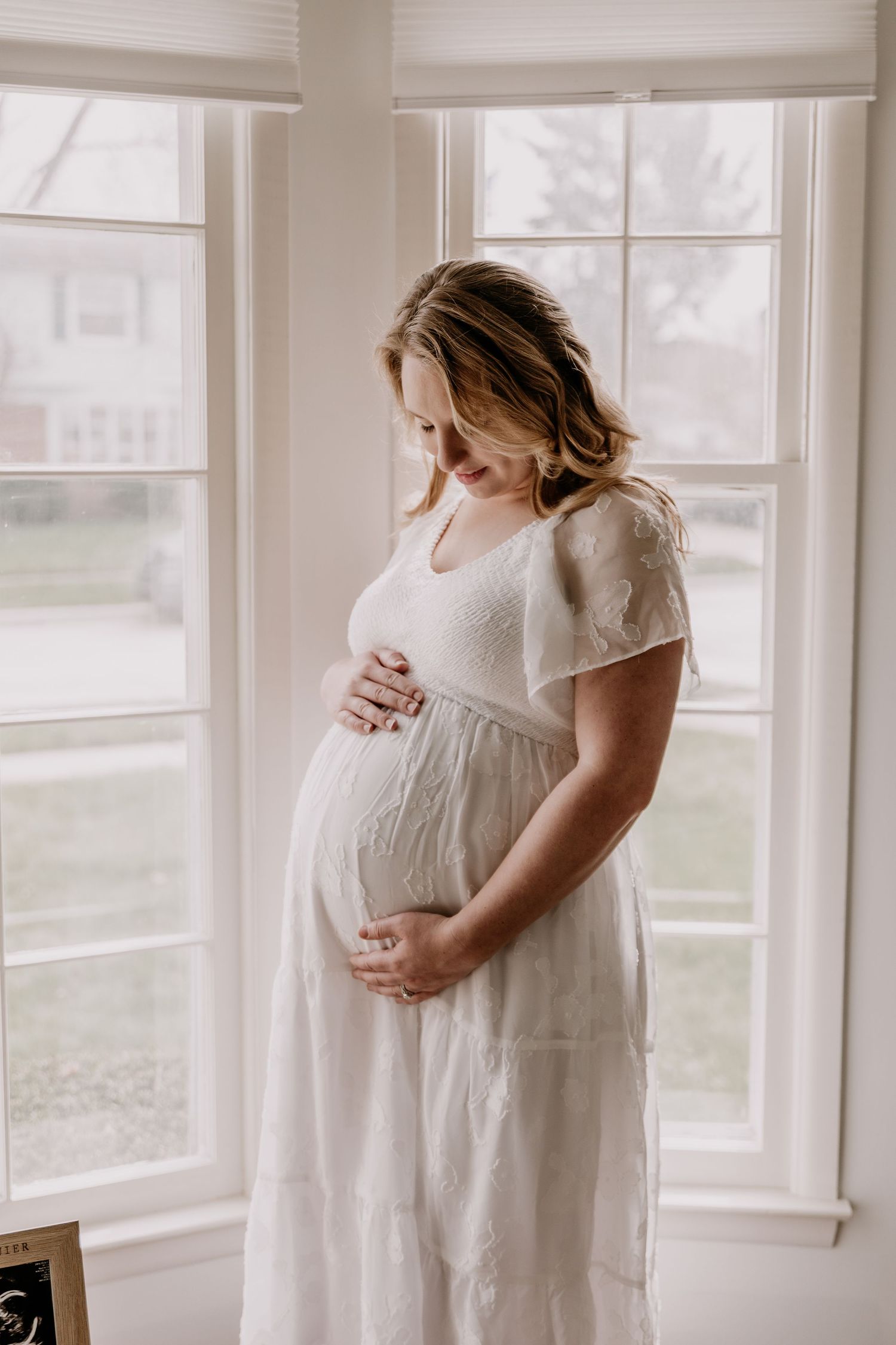 Pregnant woman in white lace dress cradling baby bump by bright windows in a serene maternity photo.
