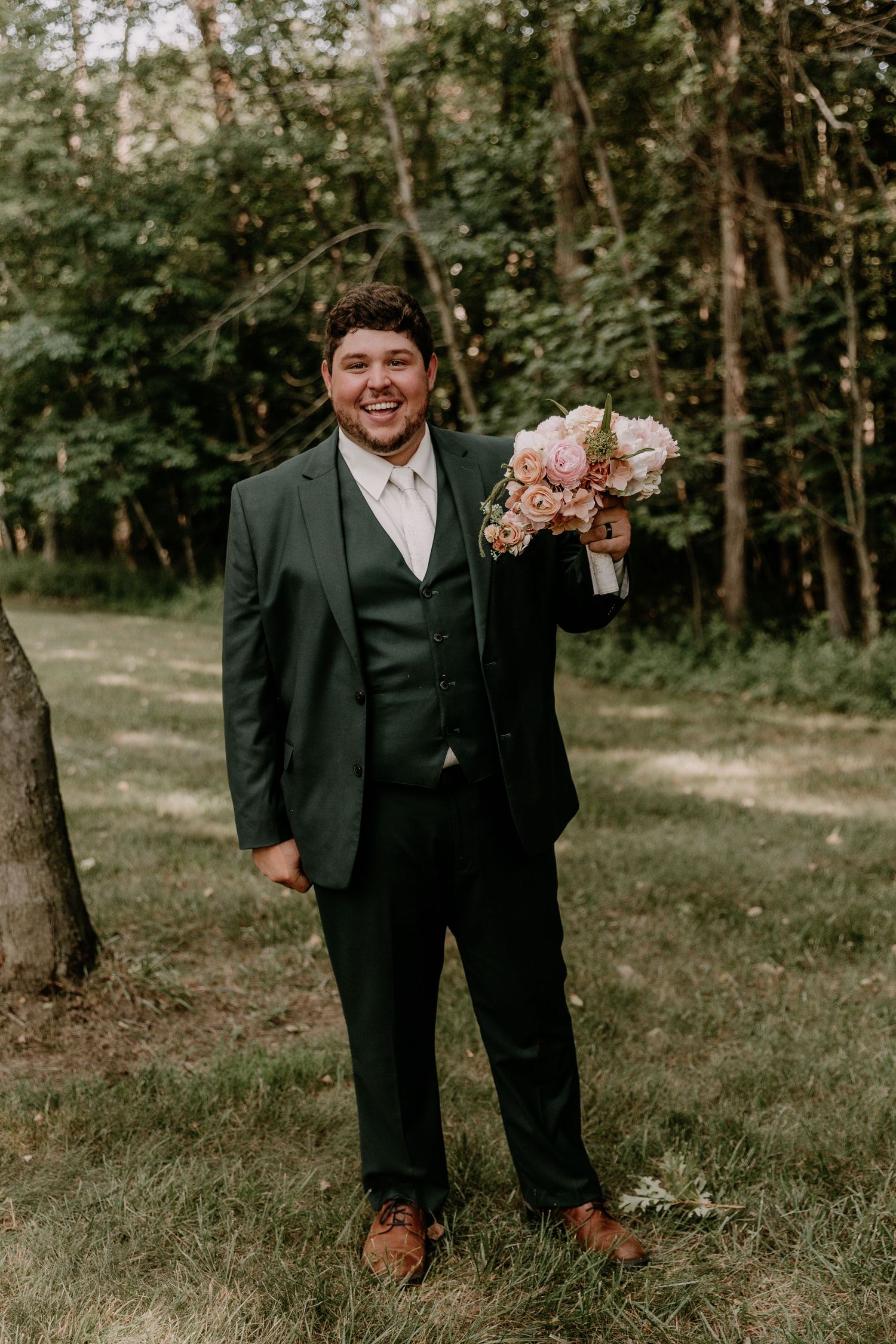 Smiling groom in dark green suit holding pink floral bouquet outdoors in a wooded garden setting.
