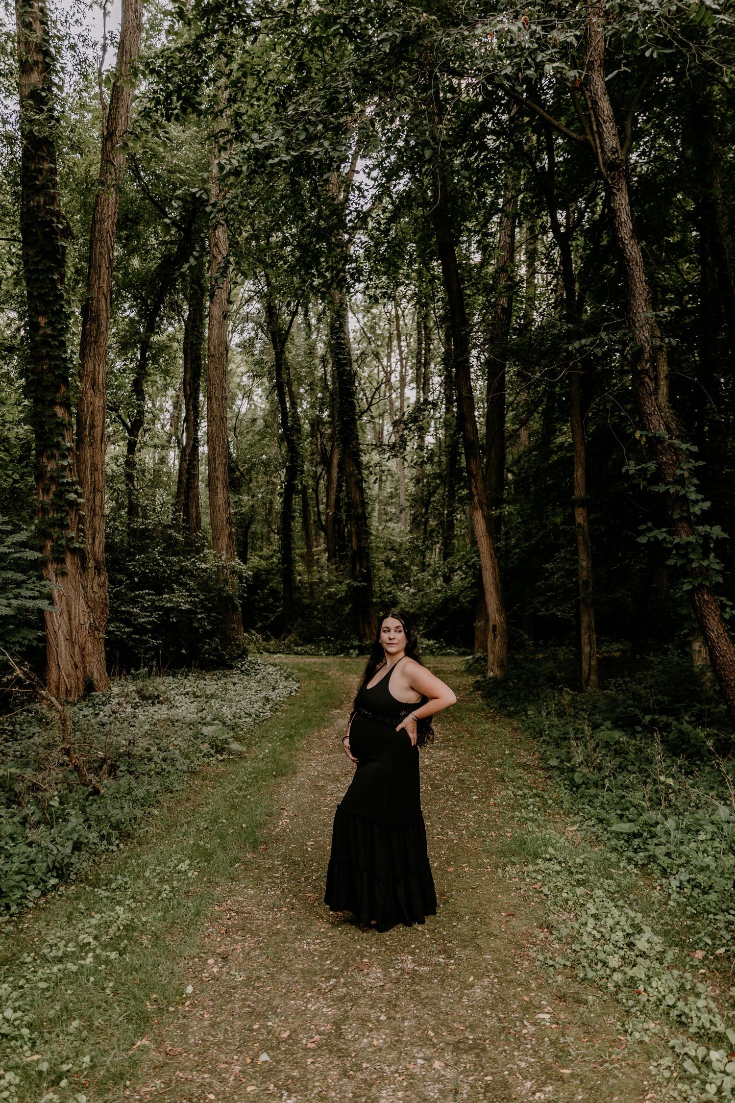 A woman in a black maxi dress stands on a forest path surrounded by tall trees and lush green foliage.