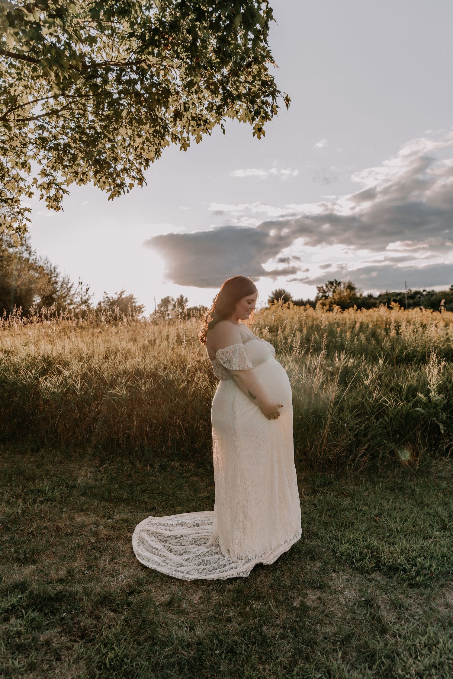 A pregnant woman in a flowing white lace dress stands in a golden field at sunset with dramatic clouds in the background.