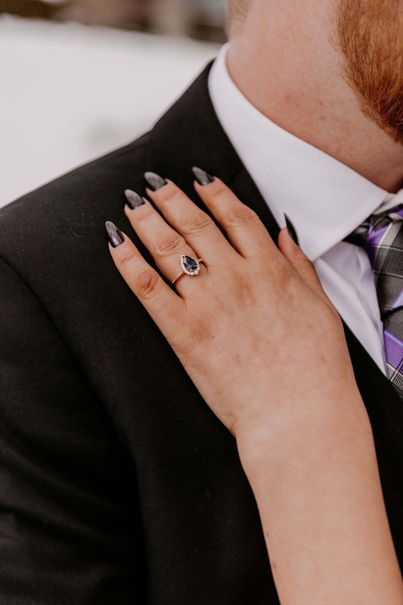 Close-up of a hand with dark nail polish and engagement ring resting on a suit lapel.