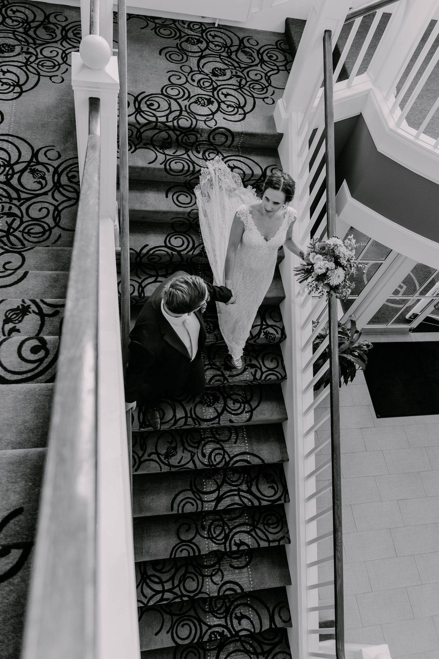 Black and white photo of couple descending ornate staircase with intricate iron railings.