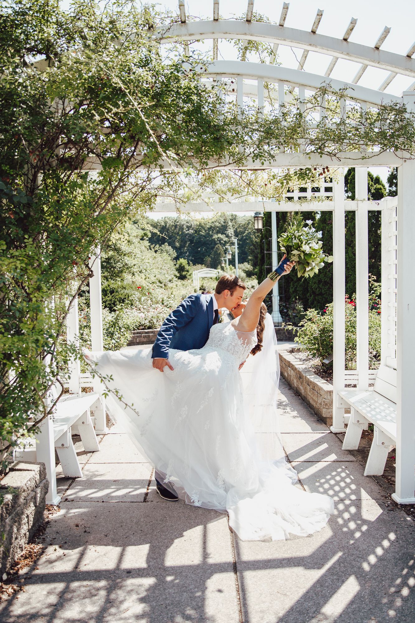 Playful wedding portrait under white pergola with bride's dress flowing in summer garden.