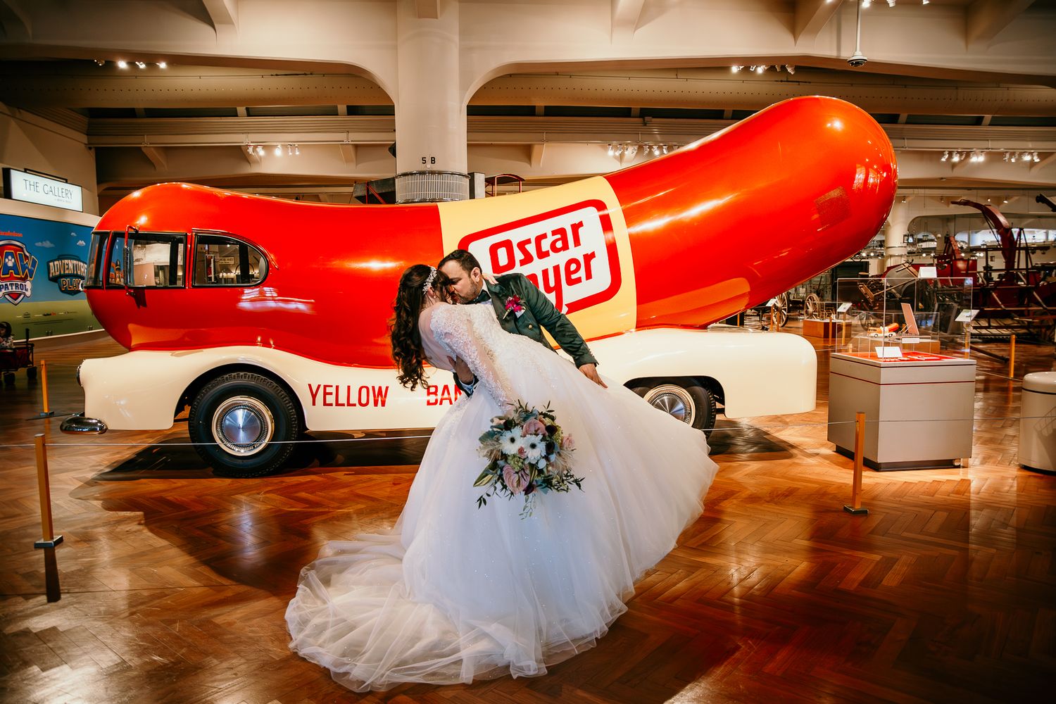 A bride in white dress posing playfully next to a giant red Oscar Mayer Wienermobile in an indoor exhibition space.