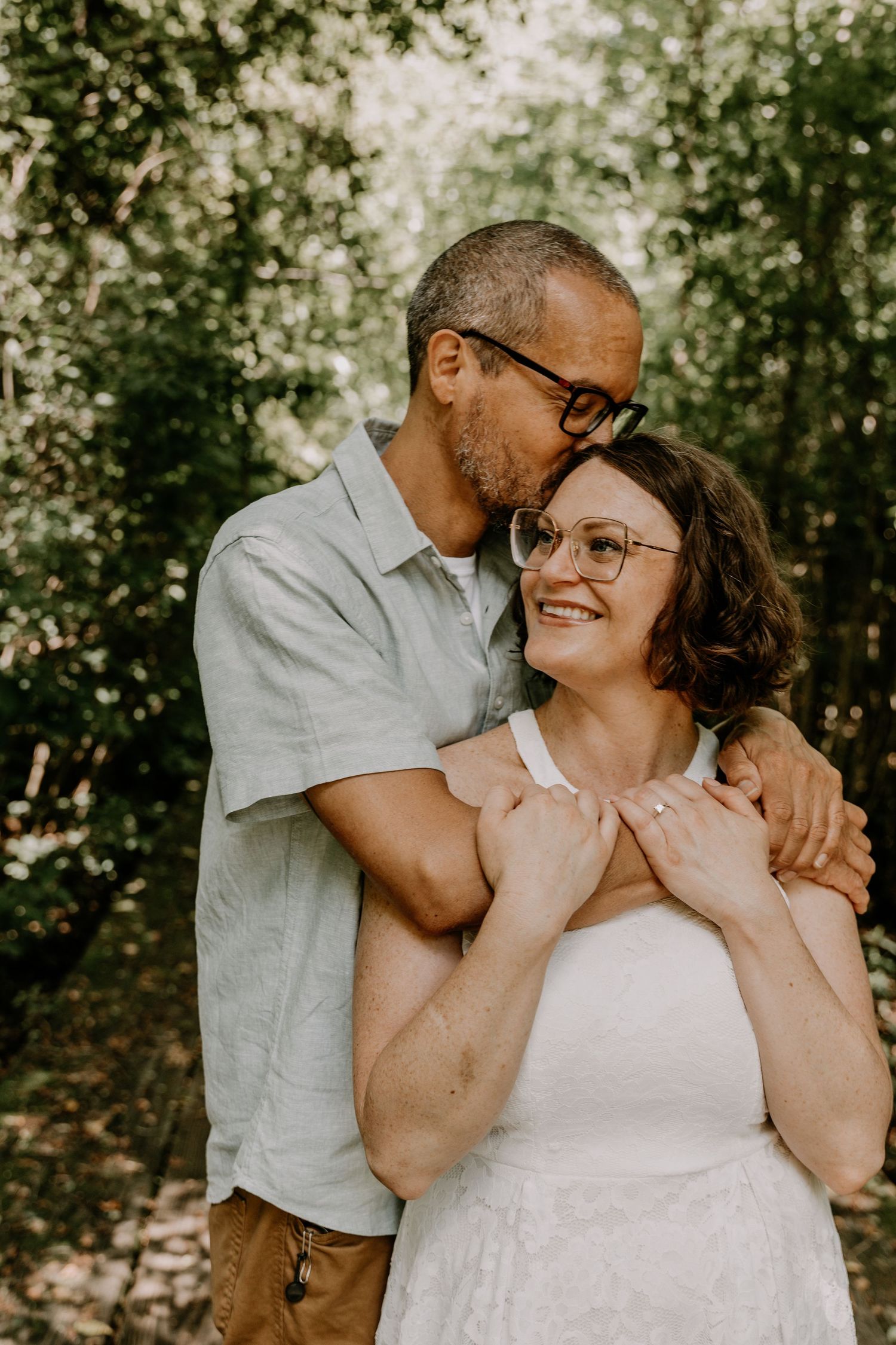 A romantic outdoor portrait of a couple embracing in dappled sunlight while wearing glasses and summer attire.