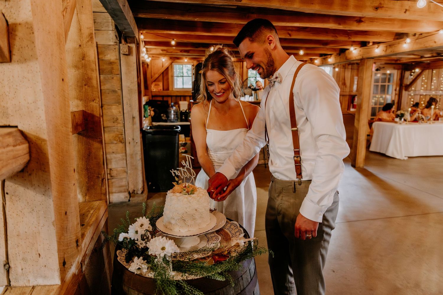 Bride and groom cut their rustic wedding cake together in a warmly lit barn reception venue.