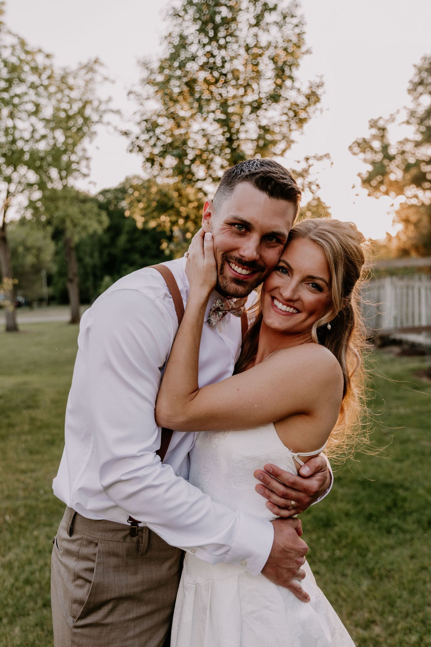 A smiling bride and groom embrace outdoors at golden hour, wearing wedding attire with suspenders and a bow tie.