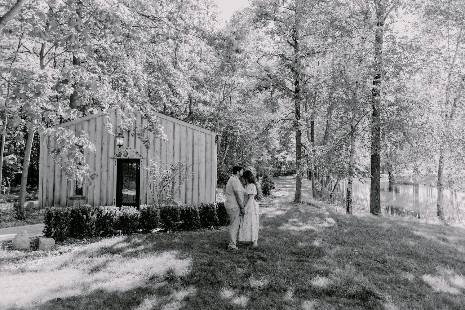 Black and white photo of a couple embracing near a rustic wooden shed surrounded by tall trees.