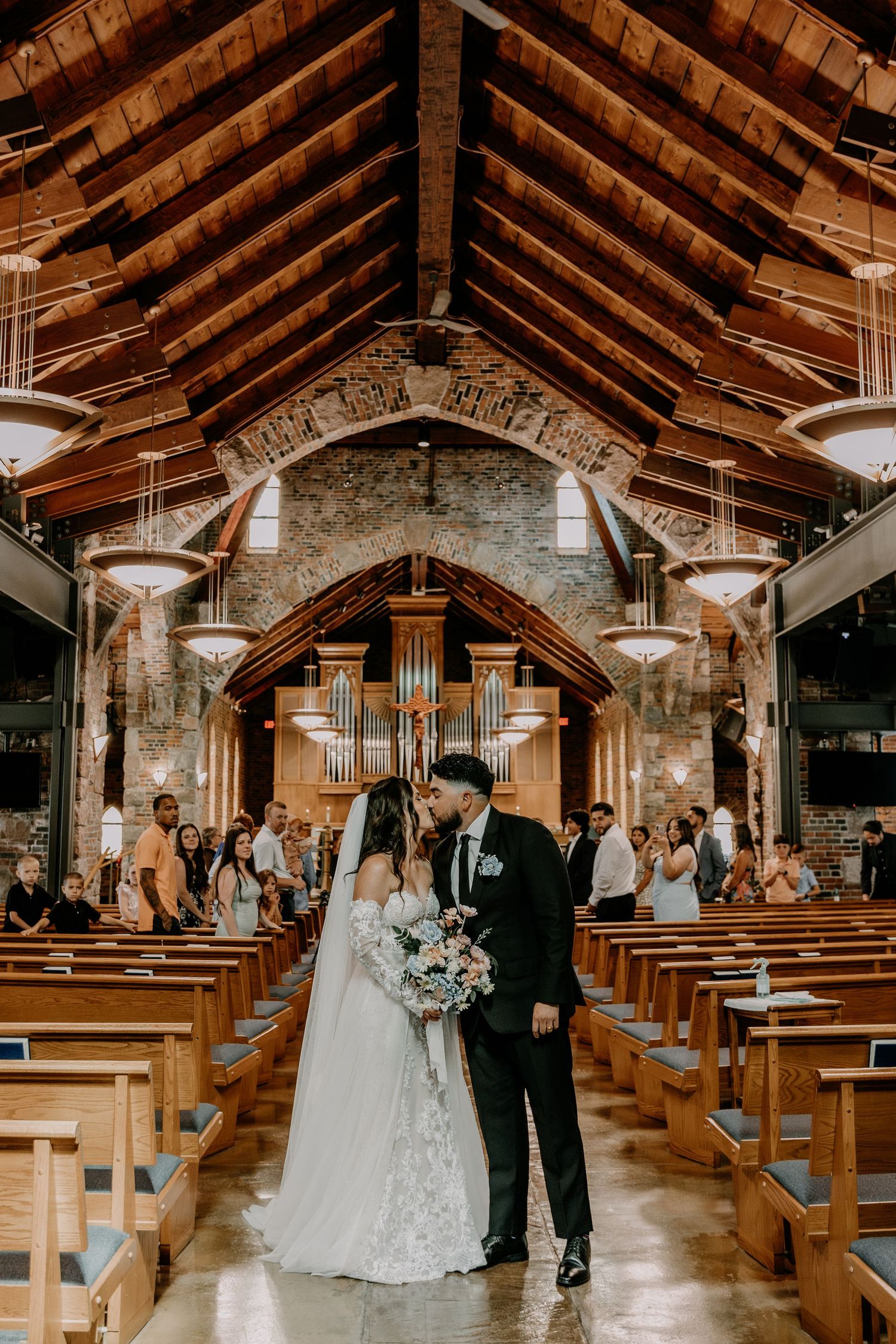 A wedding couple stands in the center aisle of a rustic stone church with exposed wooden beams and a pipe organ.