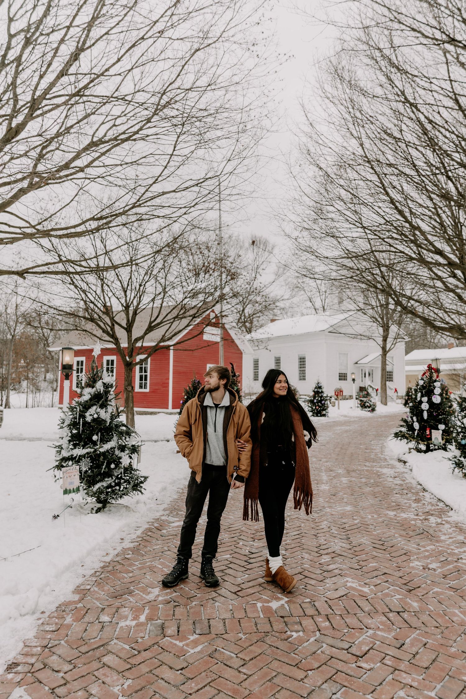 A winter scene with snow-covered brick path leading to red buildings and bare trees in the background.