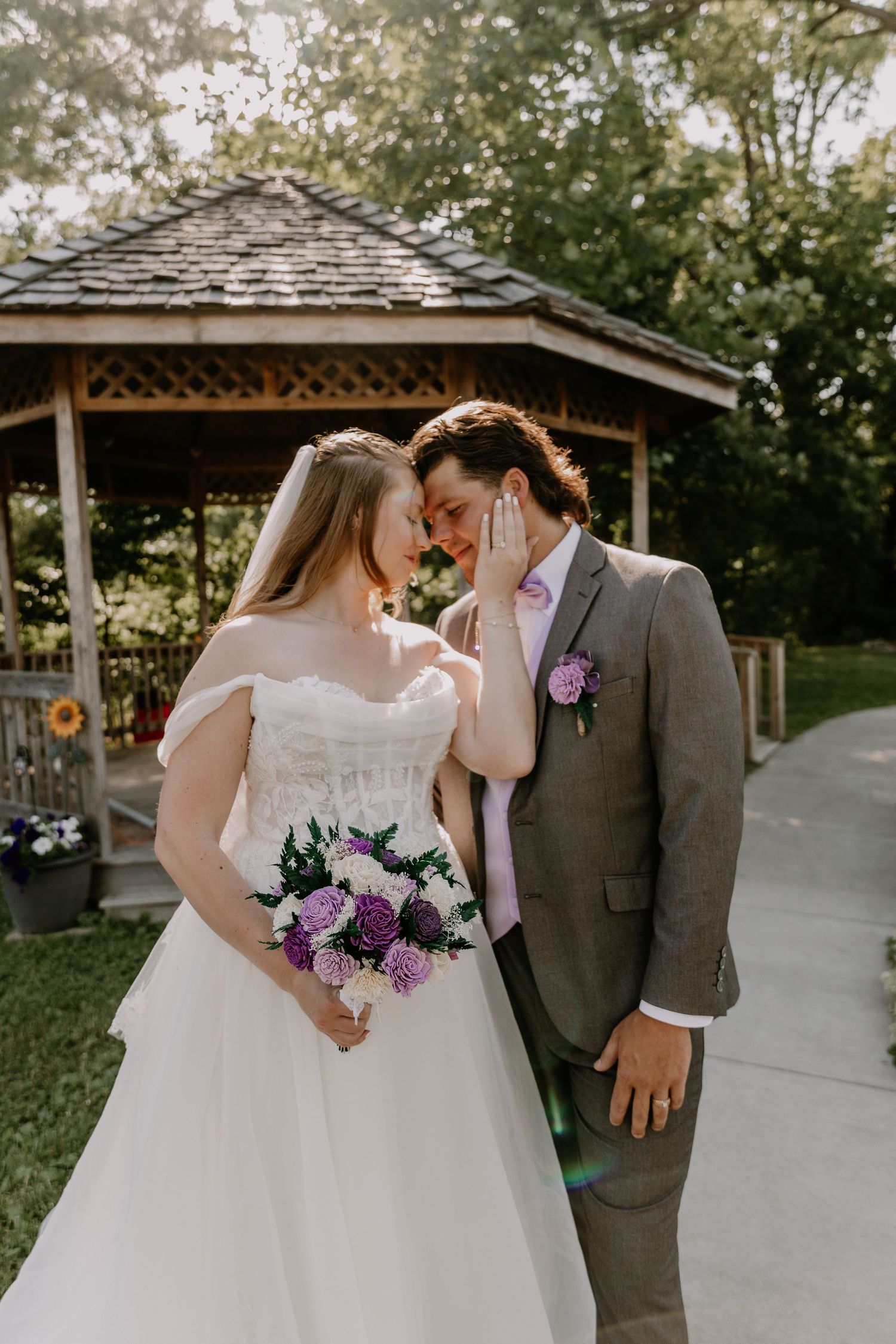Bride and groom touch foreheads near a wooden gazebo holding a purple bouquet.