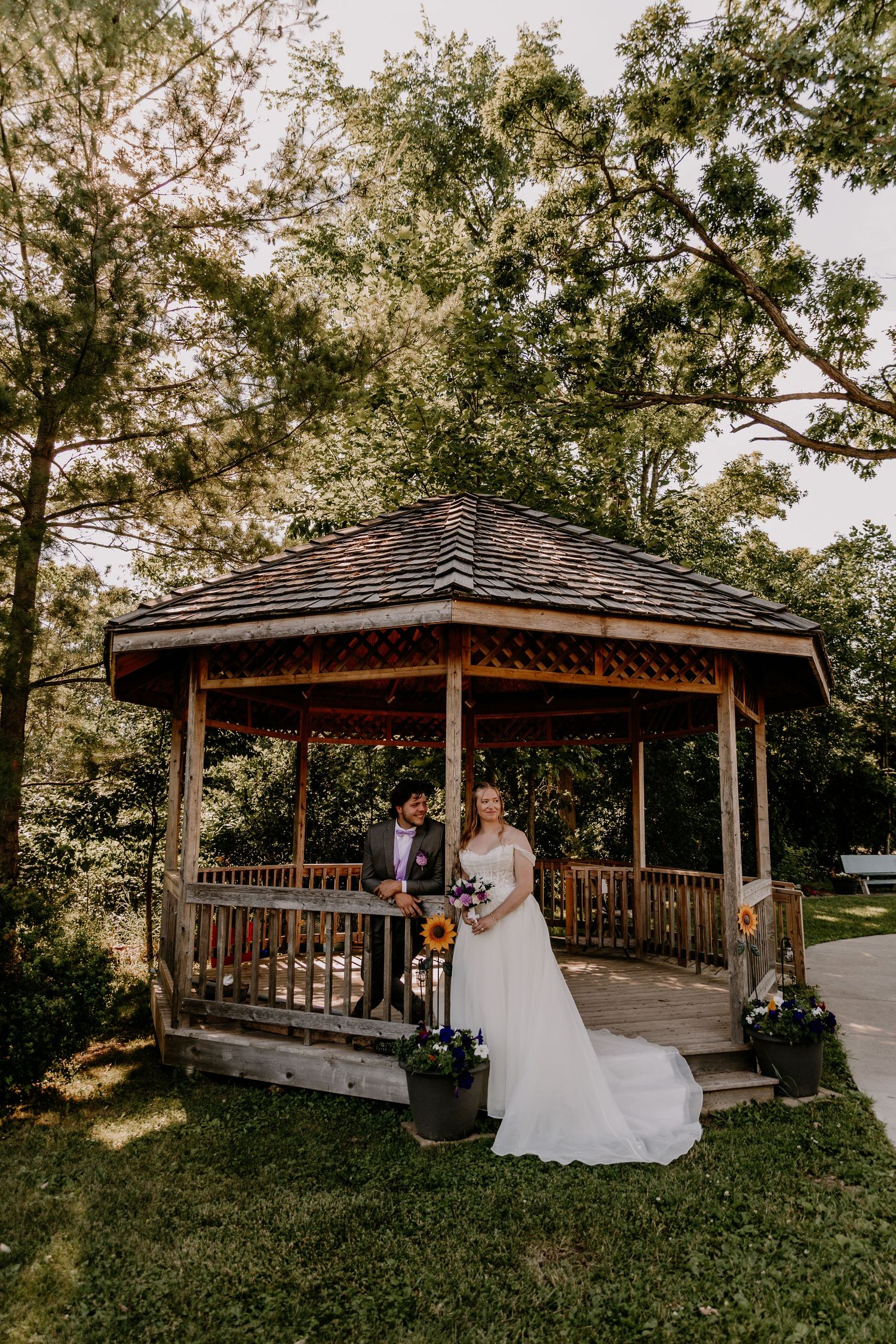 Bride and groom posing in front of a rustic wooden gazebo surrounded by lush green trees at an outdoor wedding venue.