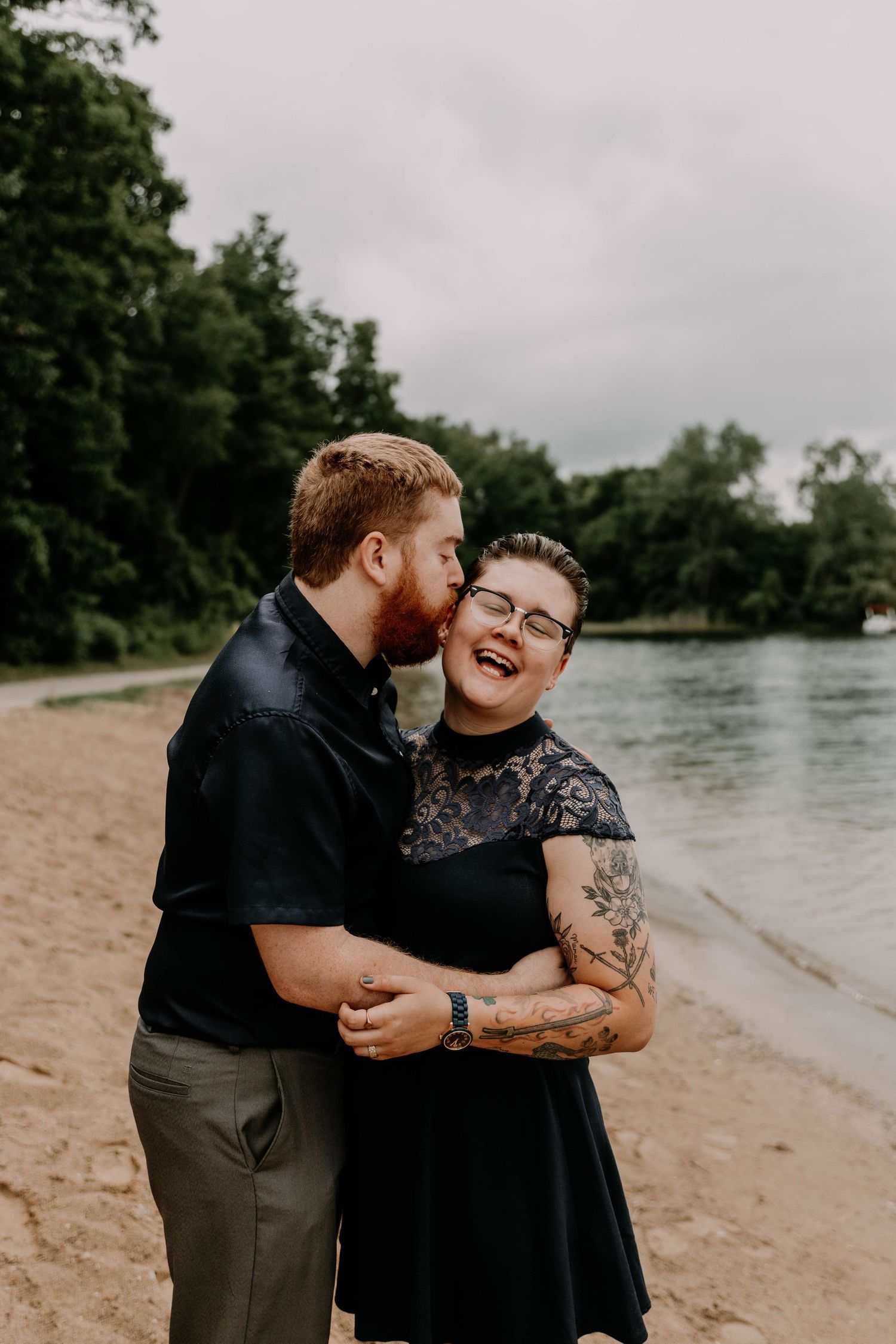 Happy couple sharing laughter and embracing on beach with water and trees in background.