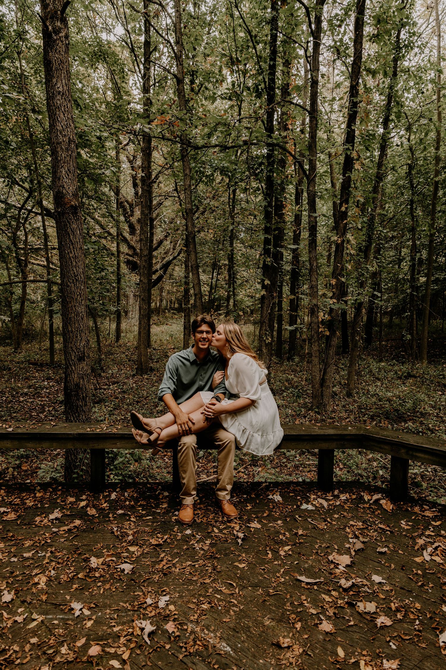 A couple shares a romantic moment on a wooden bench in a wooded forest setting.