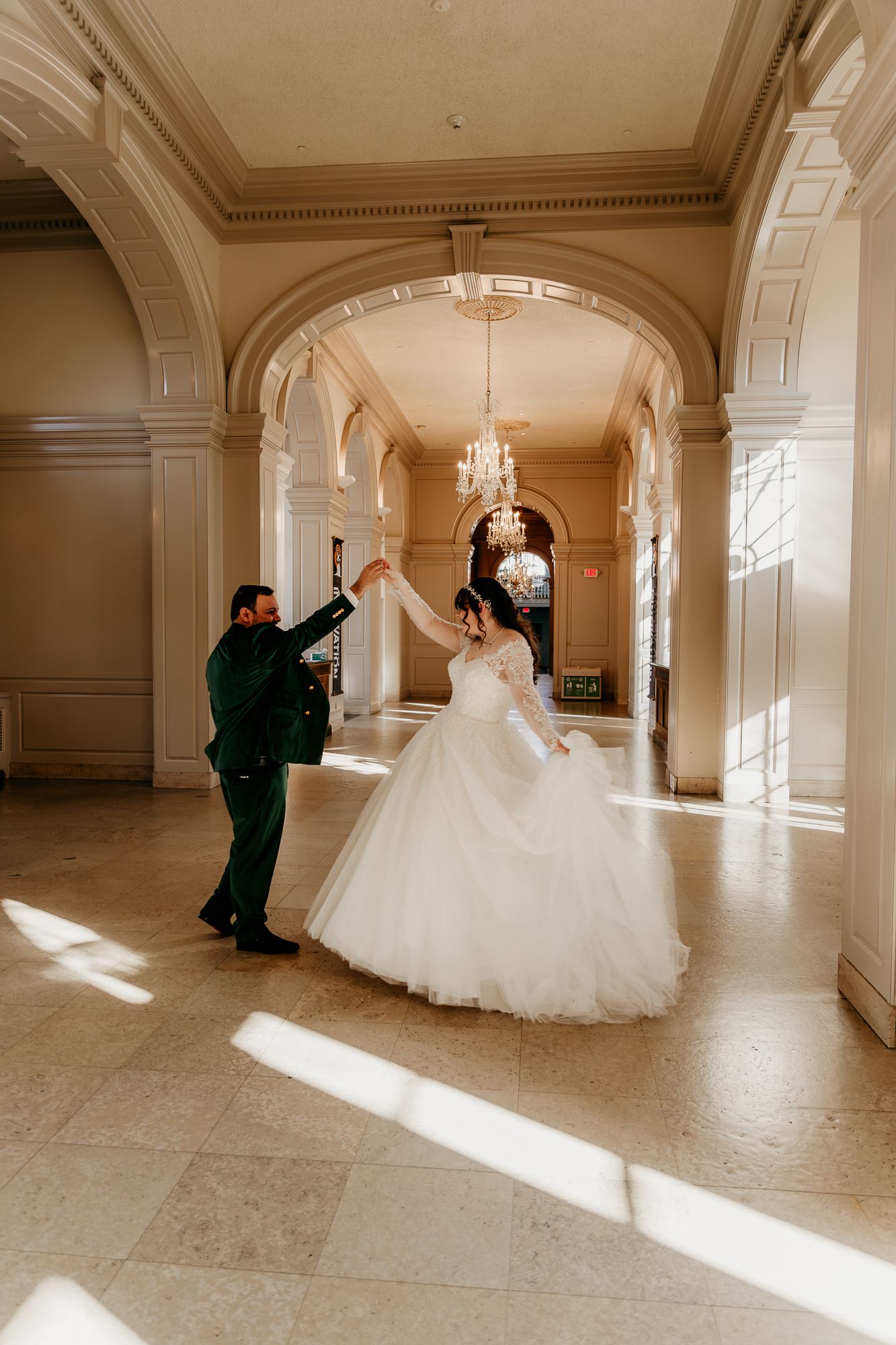 A couple twirling in a sunlit classical hallway with ornate architectural details and chandeliers.