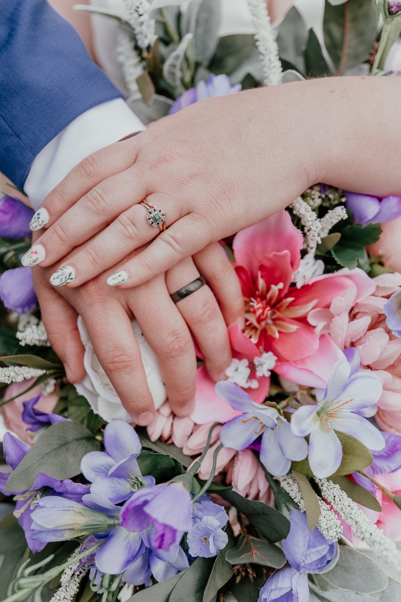 Close-up of newlyweds' hands with rings resting on a colorful bouquet of pink and purple flowers.