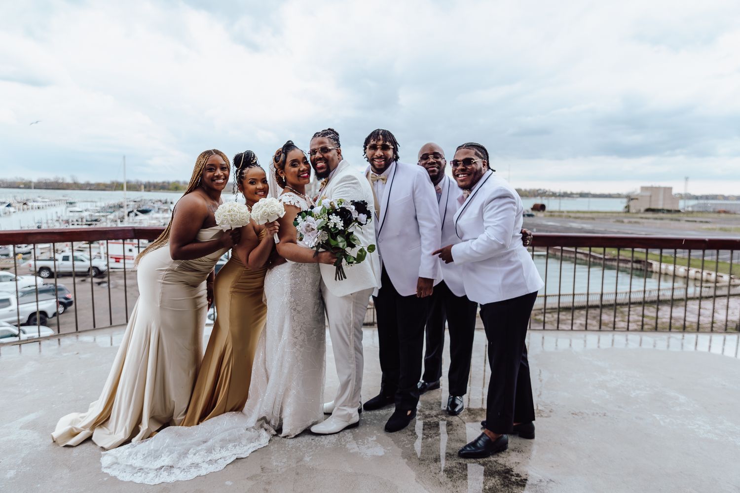 Wedding party in formal attire poses together on waterfront balcony.