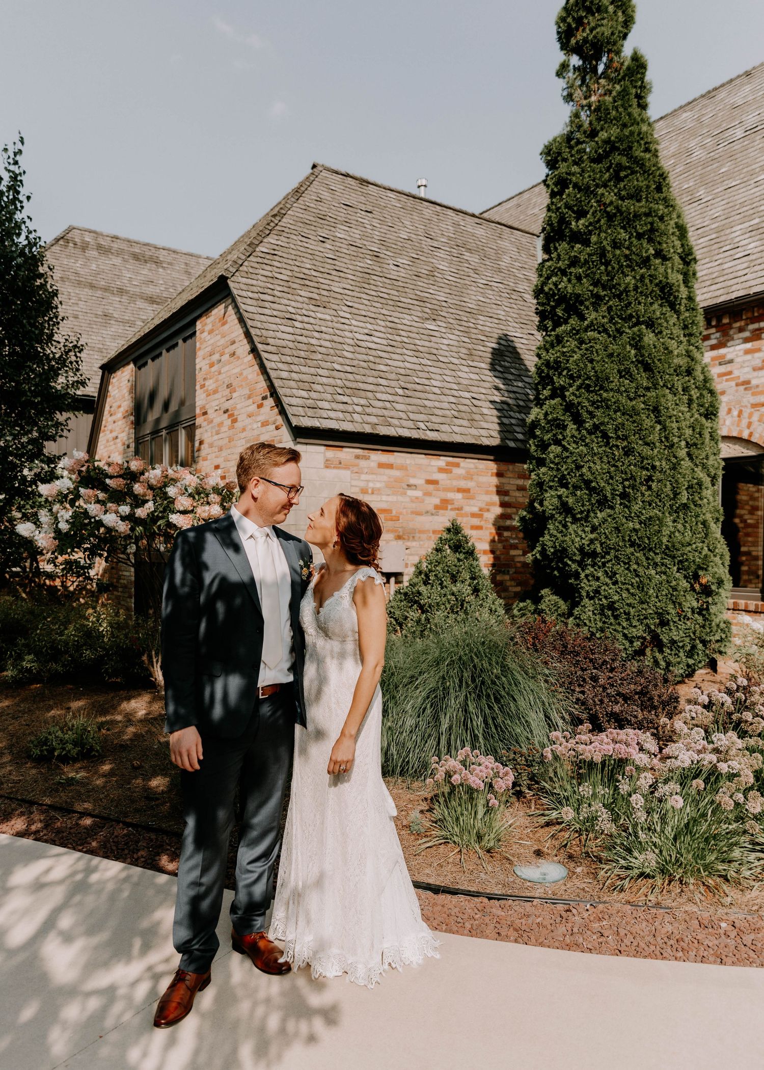 Newlyweds pose together outside a rustic stone church with tall cypress trees and lush landscaping.