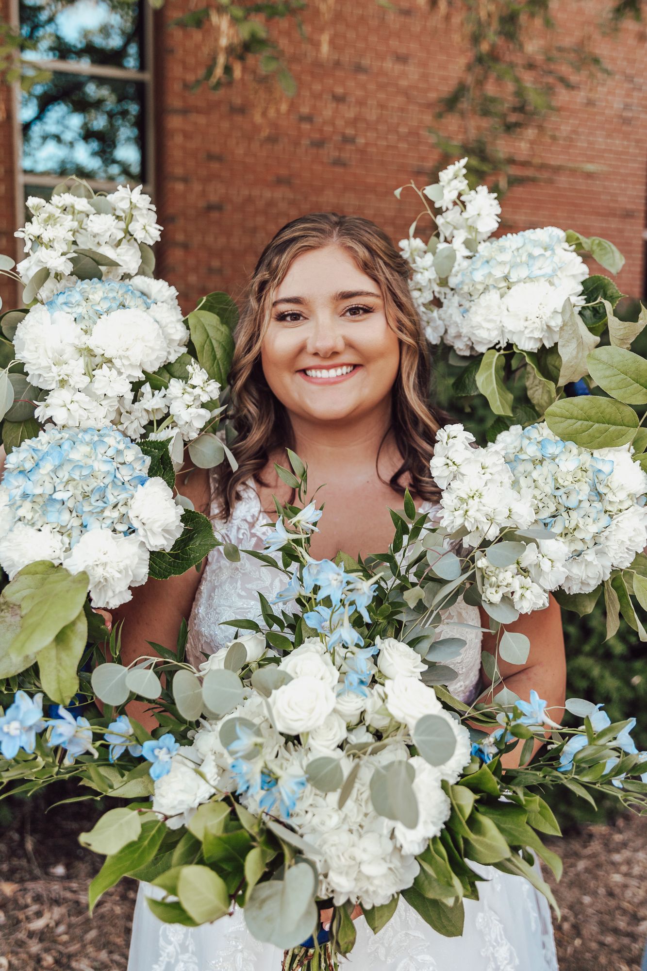 Bridal portrait featuring a large bouquet with white and light blue flowers against brick backdrop.