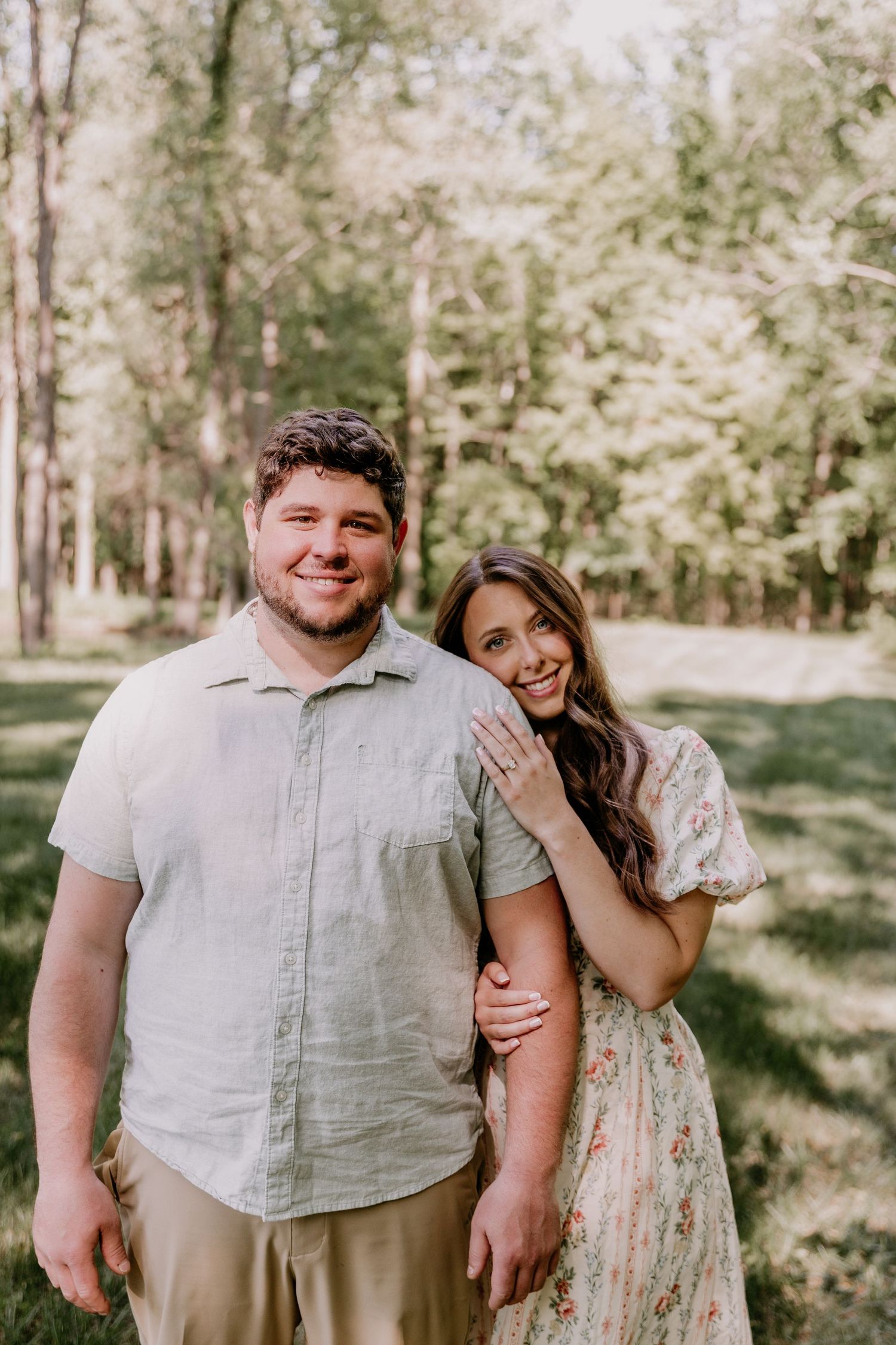 A smiling couple poses outdoors in a lush green park, woman in floral dress resting hand on mans shoulder.