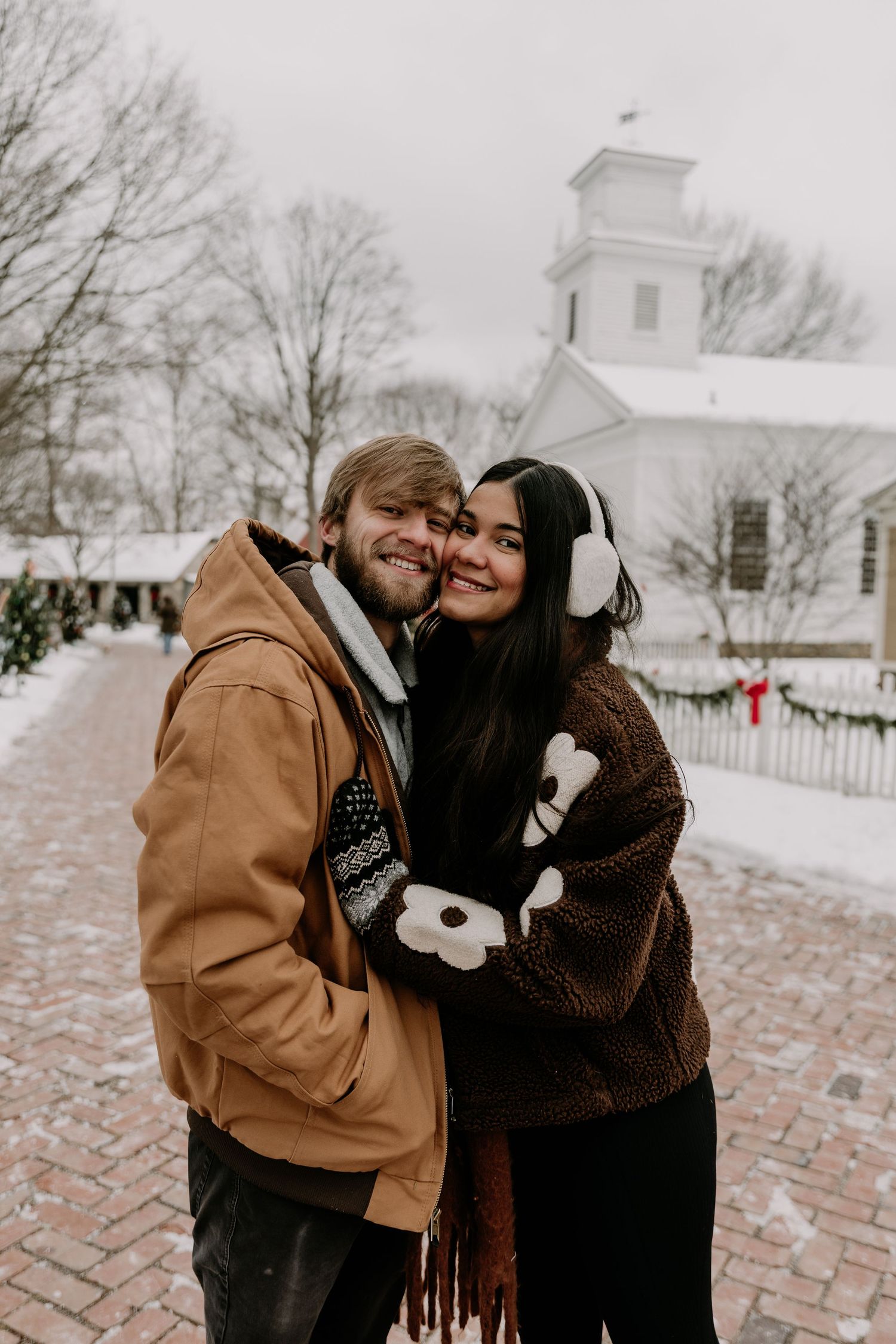 A couple shares intimate moments in the snow against a white church backdrop during winter.