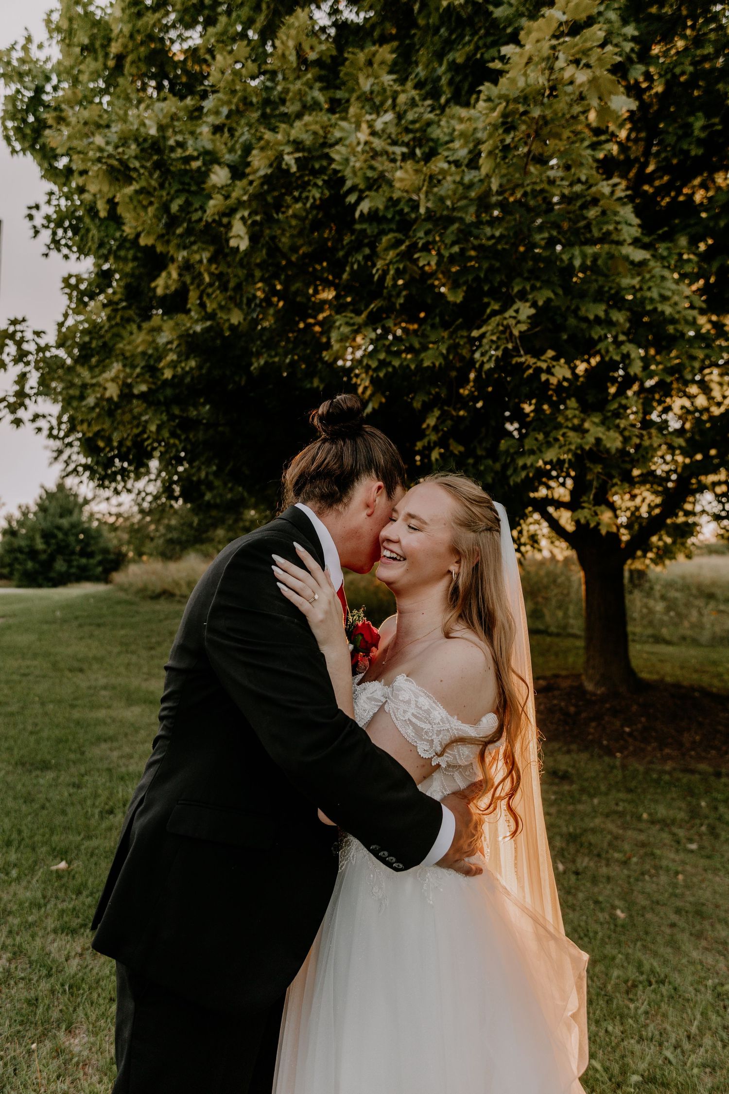 A joyful bride in a white off-shoulder gown laughs as the groom kisses her cheek outdoors near a lush green tree.