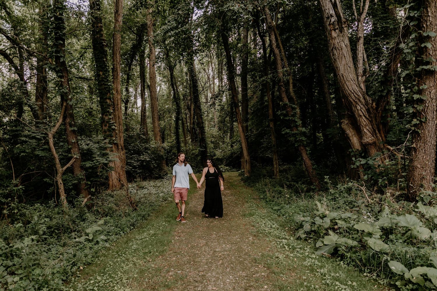 Couple holding hands walking on a forest path surrounded by lush green trees and foliage.