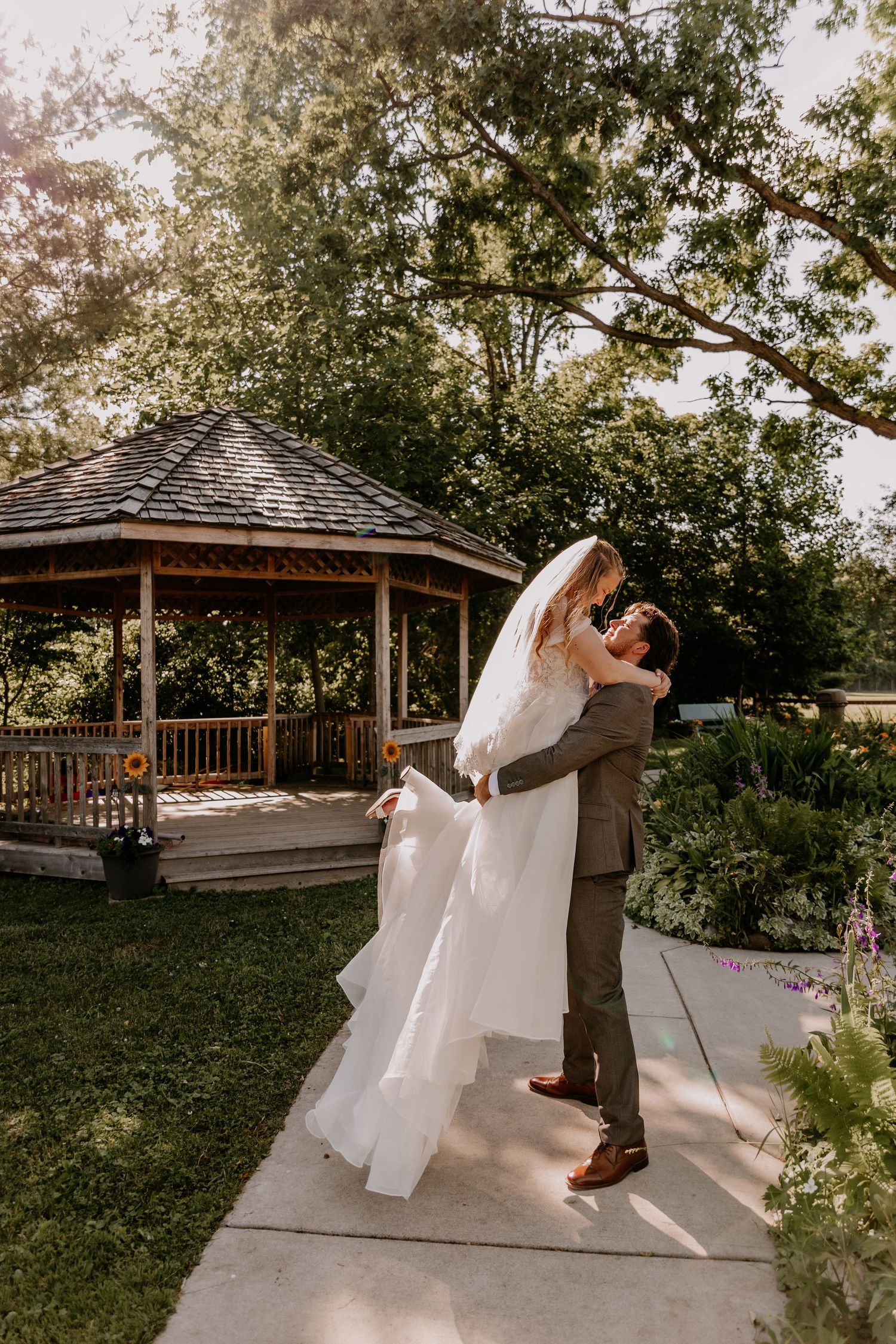 Groom lifts bride in white gown near a wooden gazebo in golden evening light.