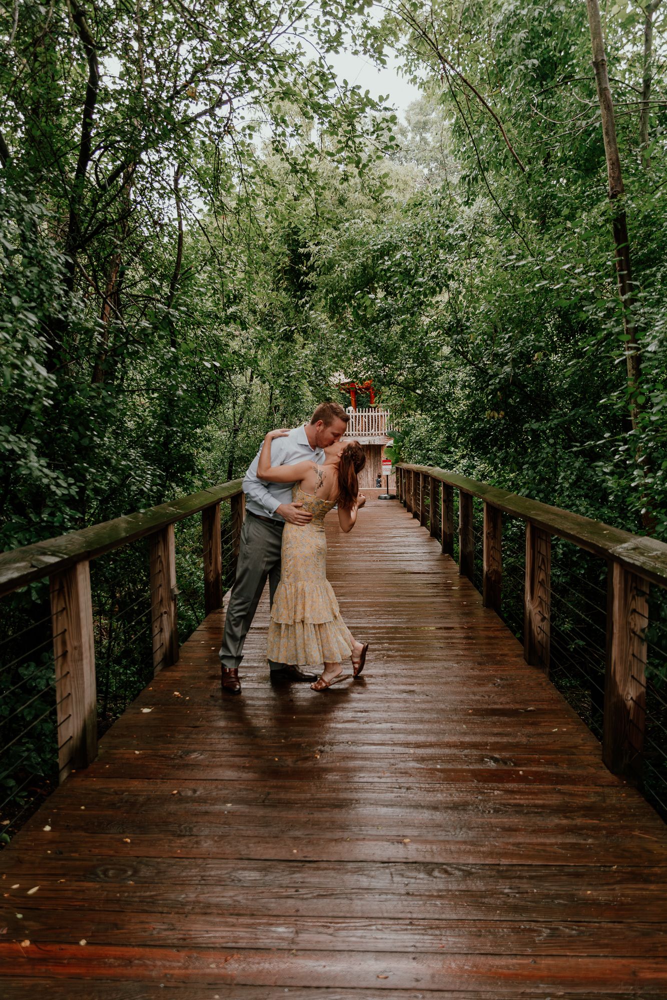 A romantic engagement photo of a couple dancing on a wooden boardwalk surrounded by green trees.