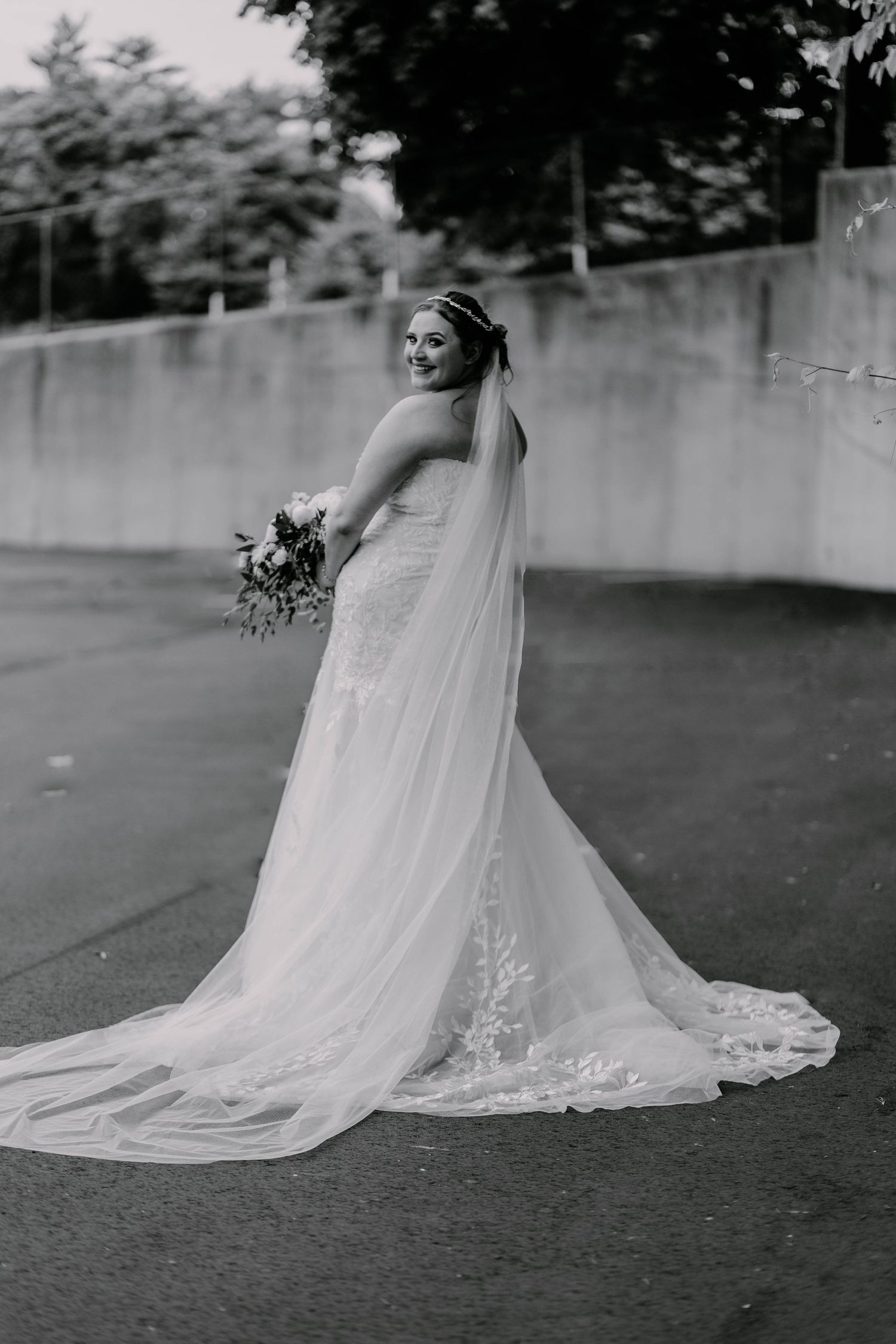 A bride in an elegant lace wedding dress with a flowing veil poses on a concrete path for a black and white portrait.