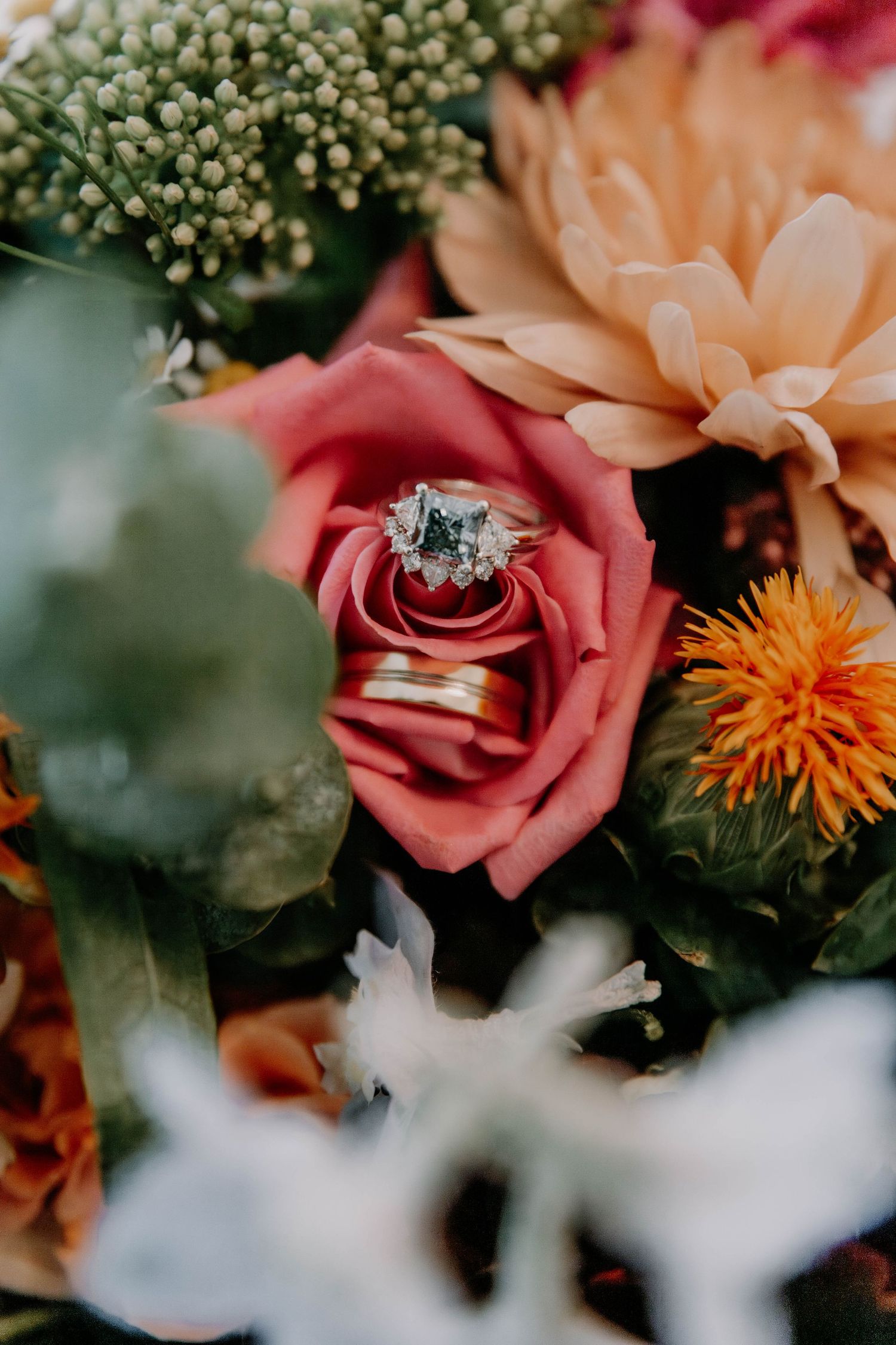 Wedding rings nestled in a coral rose surrounded by colorful flowers and greenery.