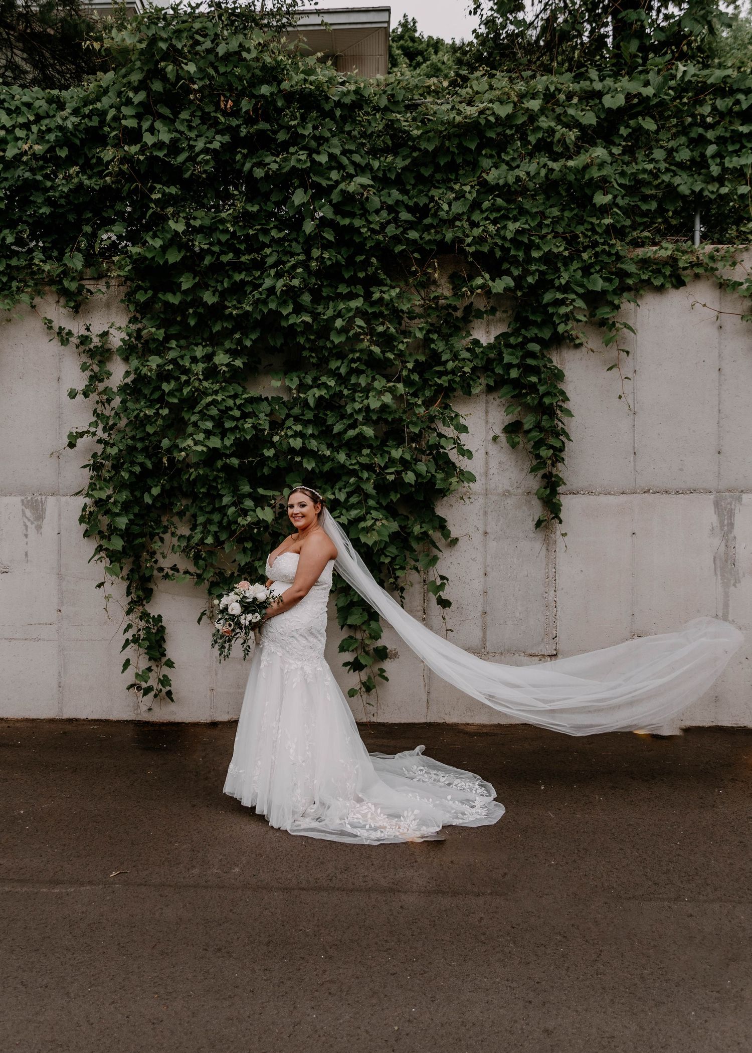 Bride's dress train flows dramatically against concrete and ivy backdrop.