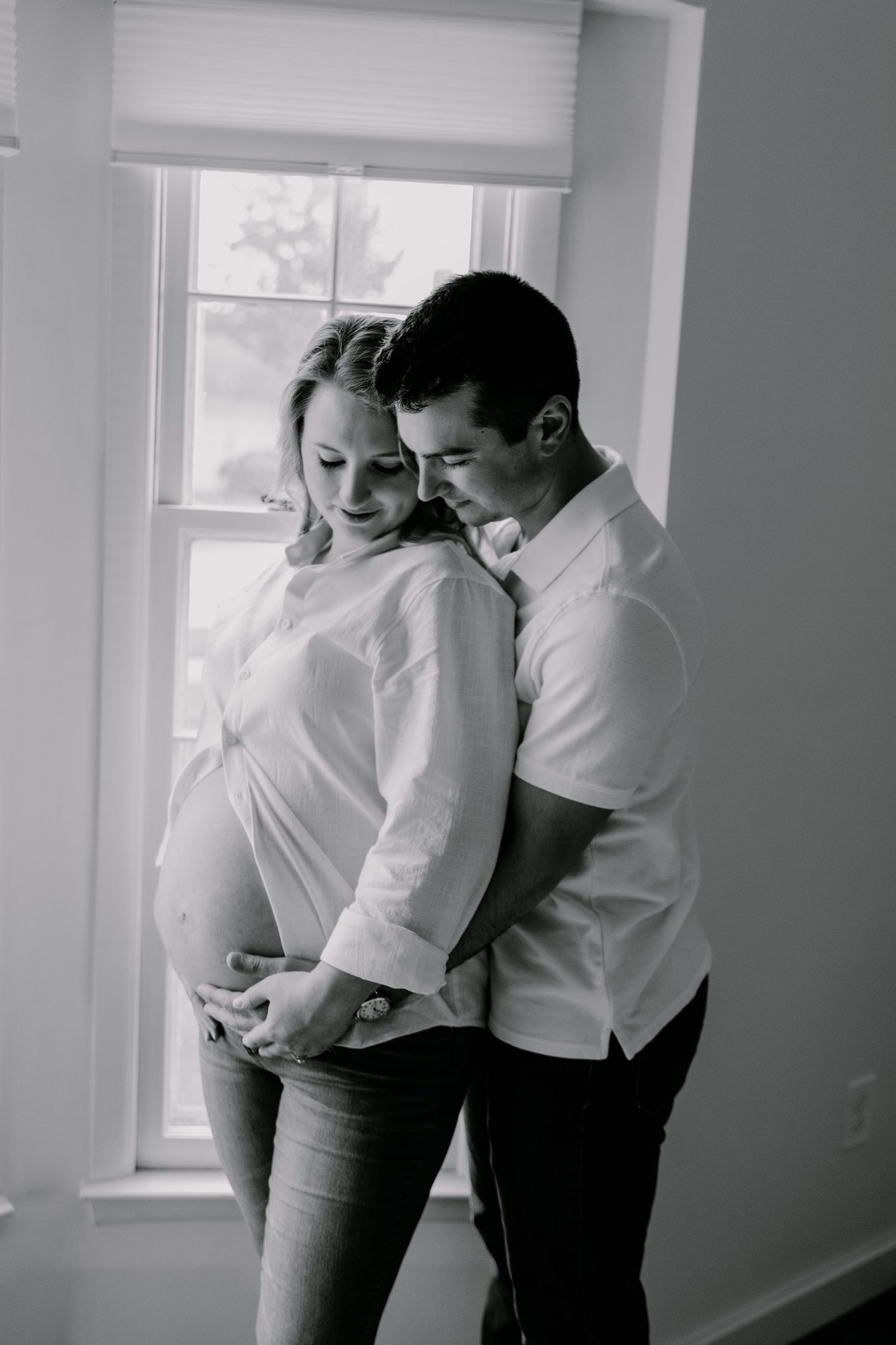 Black and white maternity photo of a couple embracing by a window, man holding pregnant woman's belly indoors.