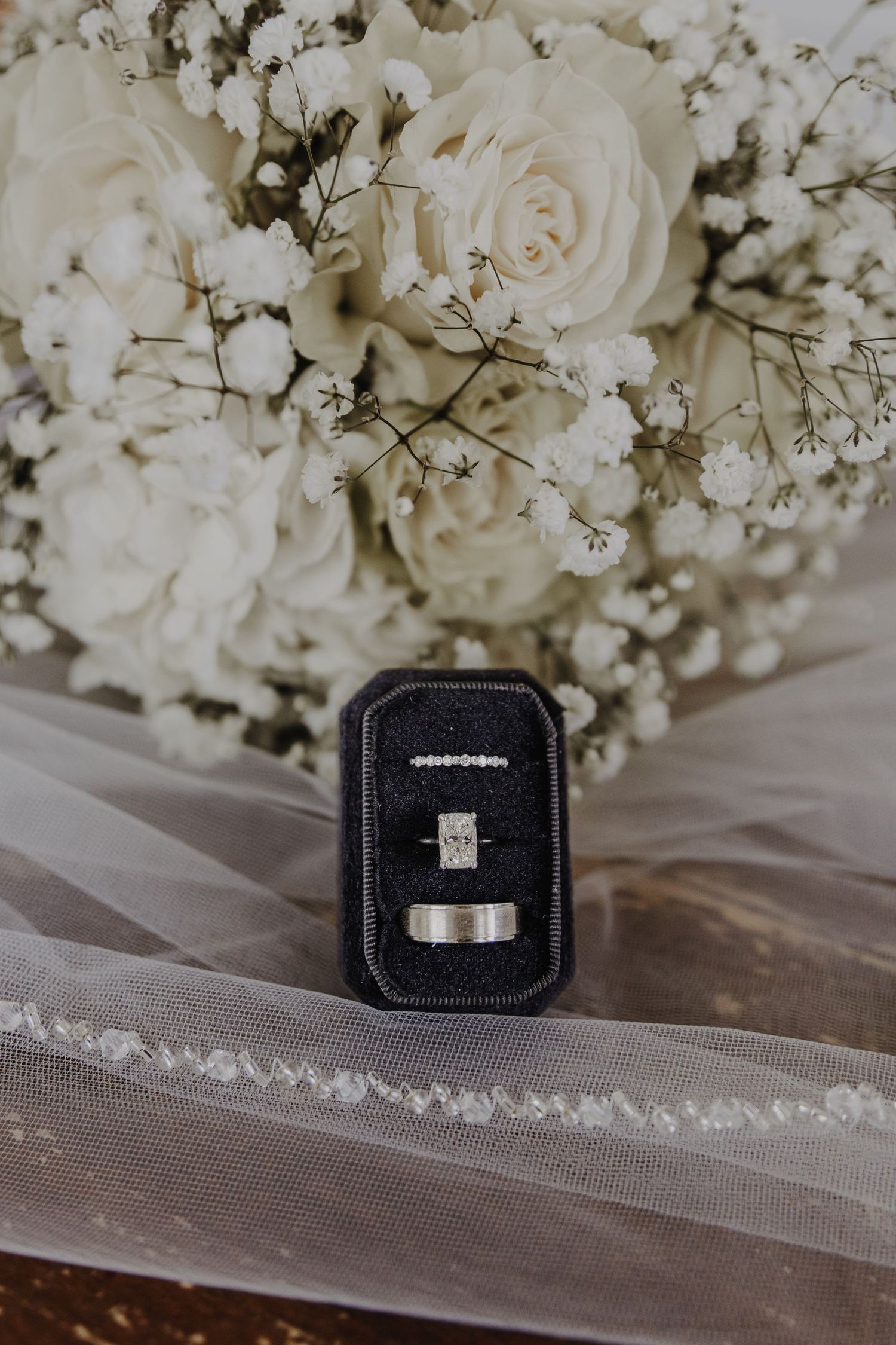 Wedding rings displayed in a black velvet box with white roses and baby's breath bouquet in the background.
