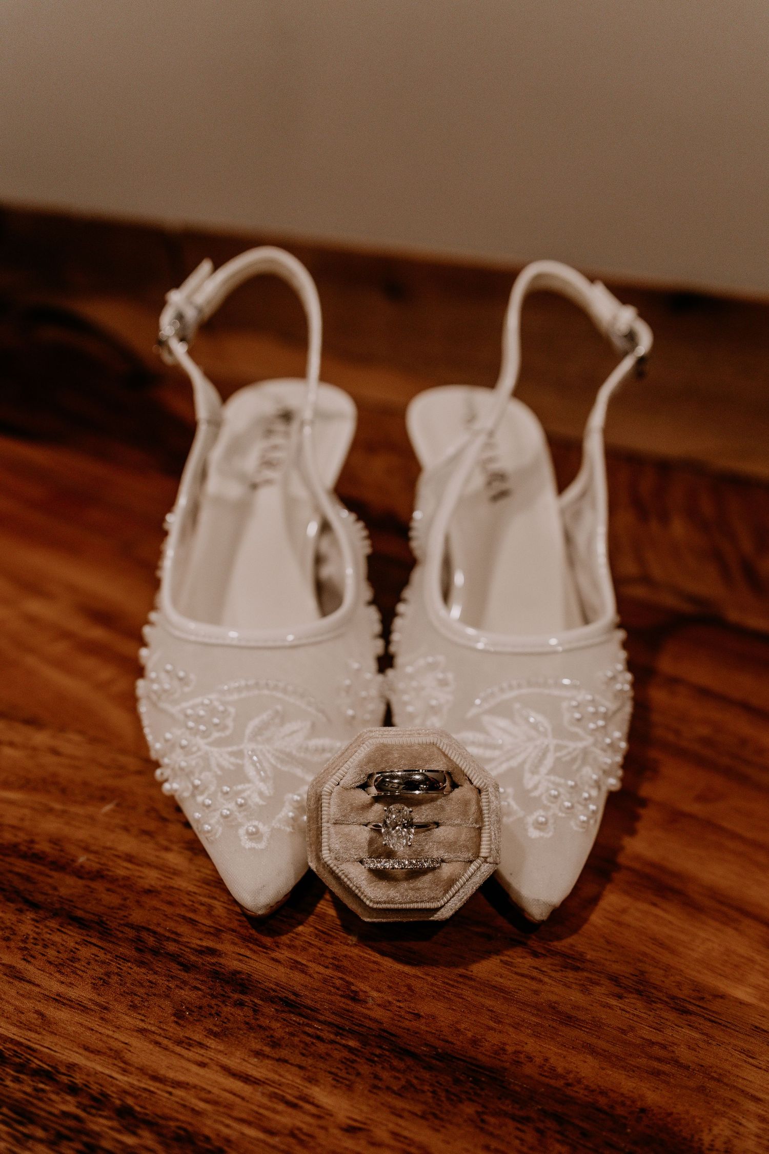 White beaded bridal slingback flats with floral embroidery displayed on wood surface with wedding rings in velvet box.