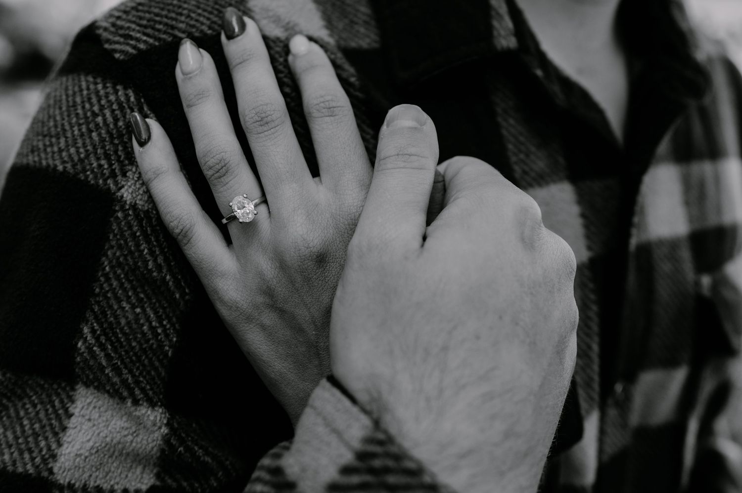 Black and white photo of a woman's hand with an engagement ring resting on a man's plaid flannel shirt.