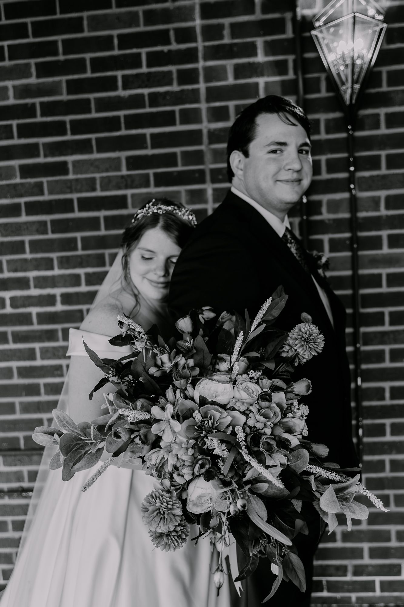 Wedding couple poses with floral bouquets against brick wall background.