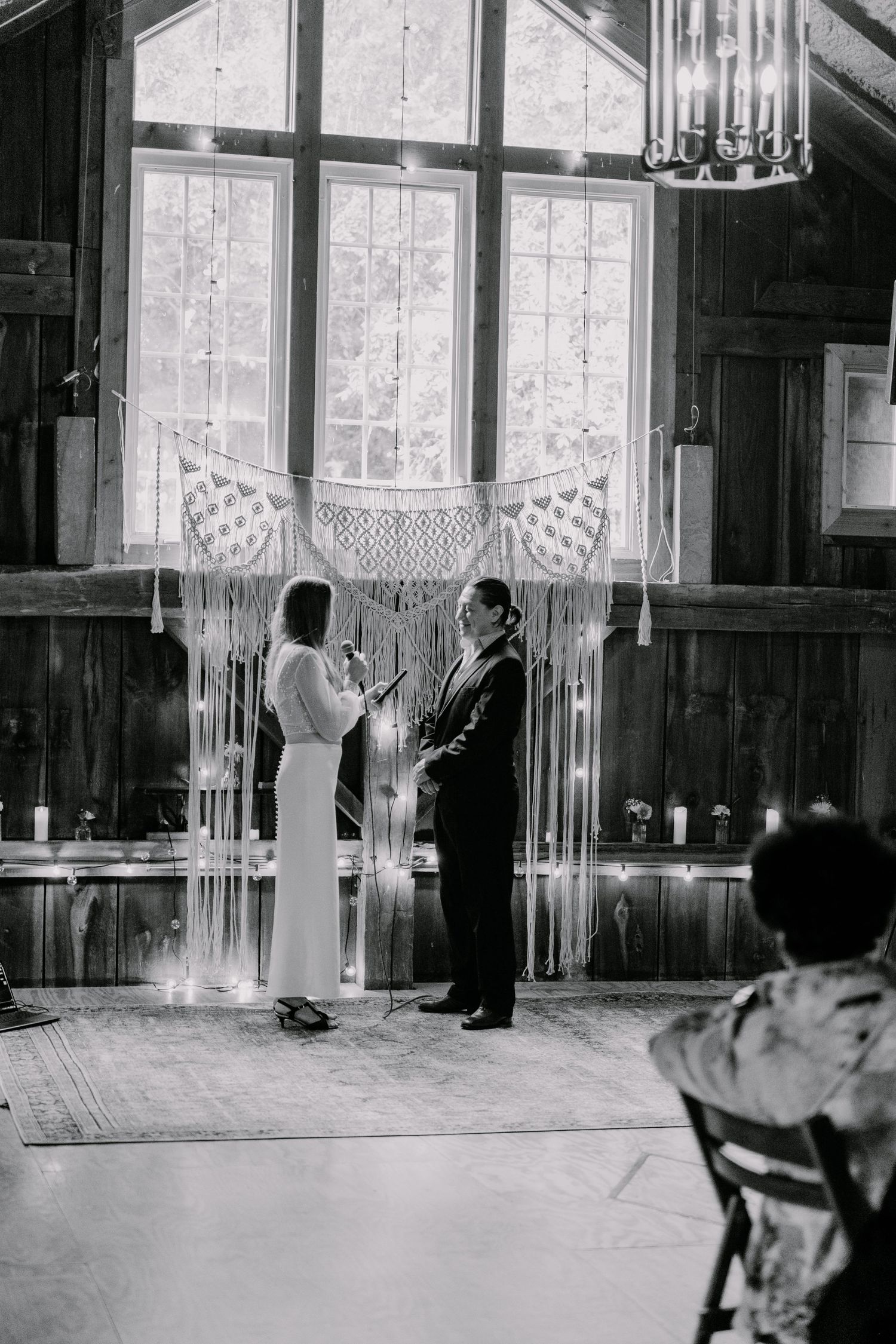 Black and white photo of indoor wedding ceremony with macrame backdrop and natural lighting.