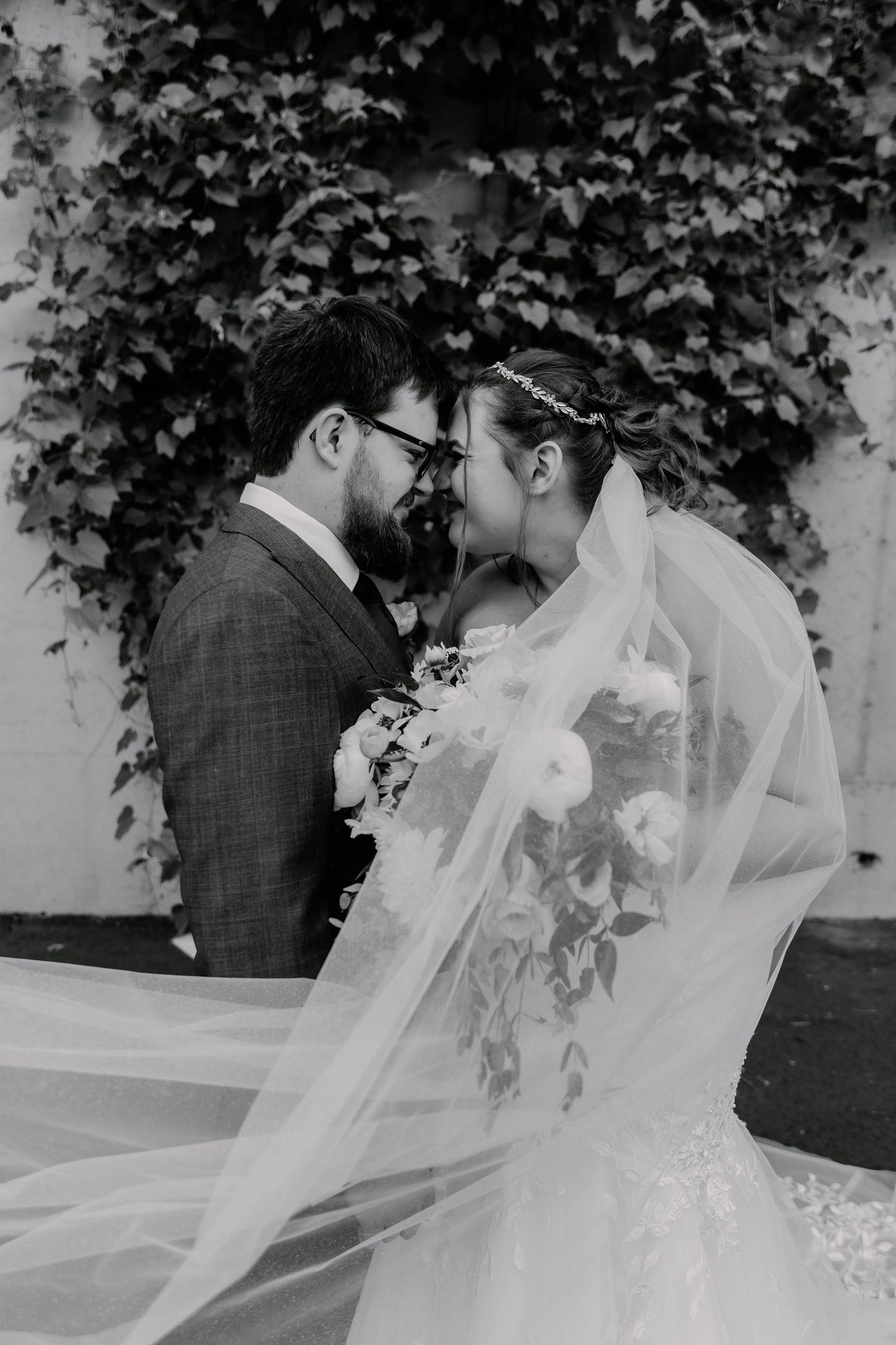 Romantic black and white candid wedding portraits of a couple with flowing veil against an ivy backdrop.