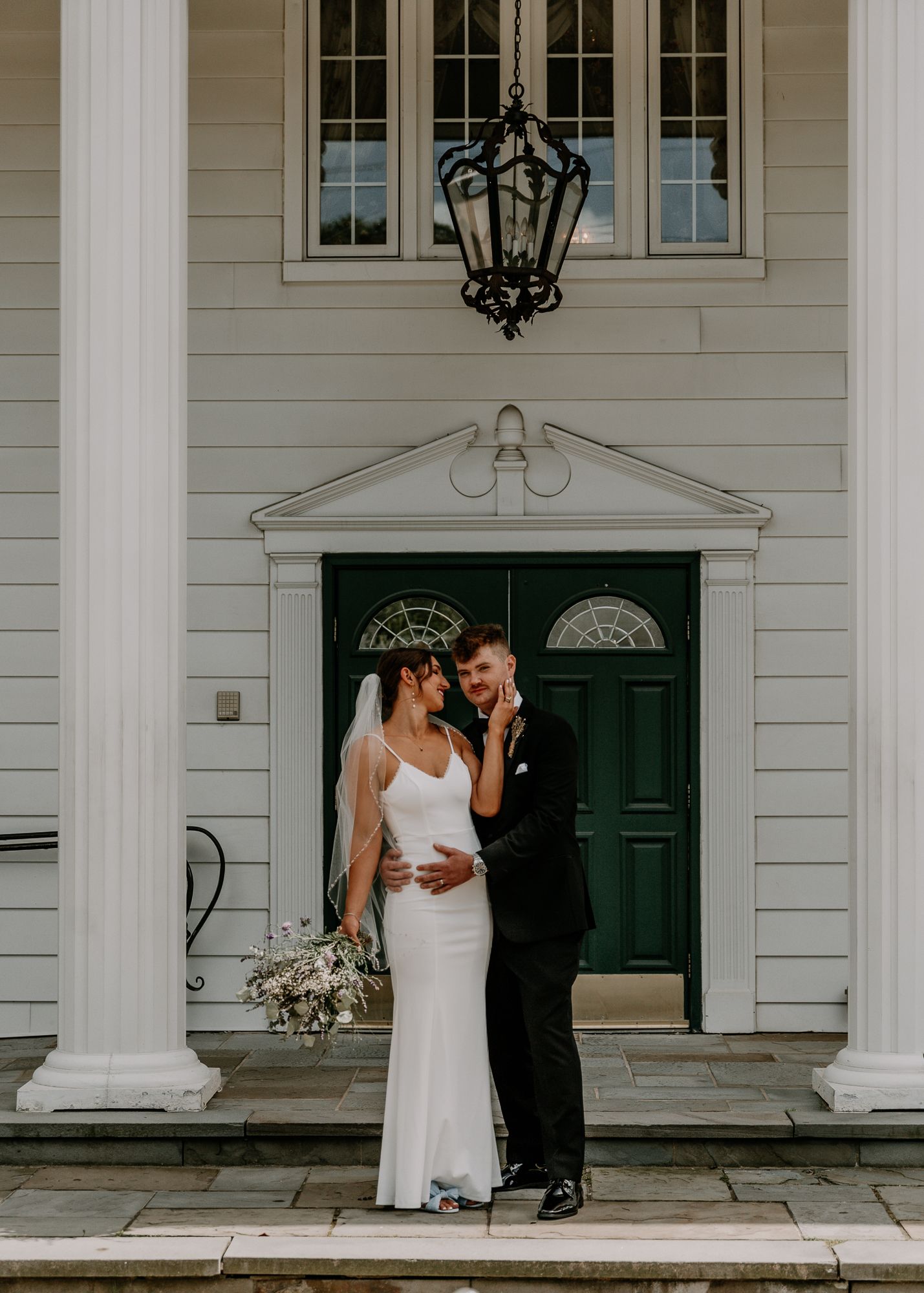 A wedding couple shares an intimate moment in front of an elegant white house with green door and hanging lantern.