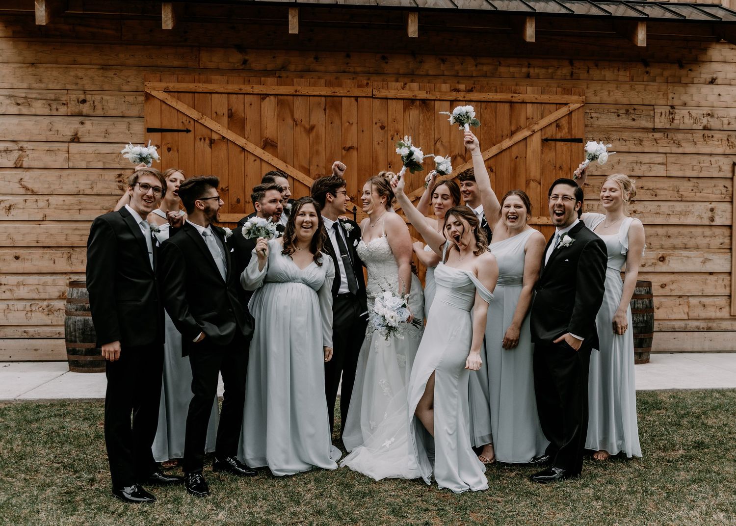 Wedding party celebrates with raised arms in front of rustic wooden barn venue during outdoor photo session.