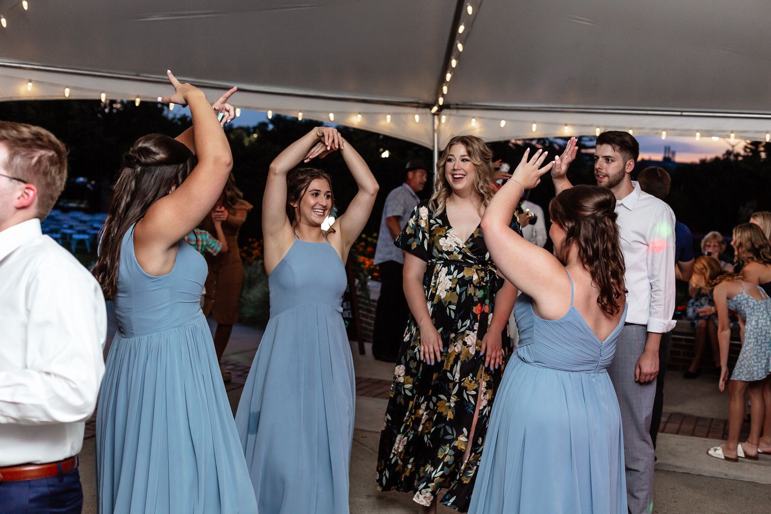 Wedding guests dancing and celebrating under tent with string lights at reception party.