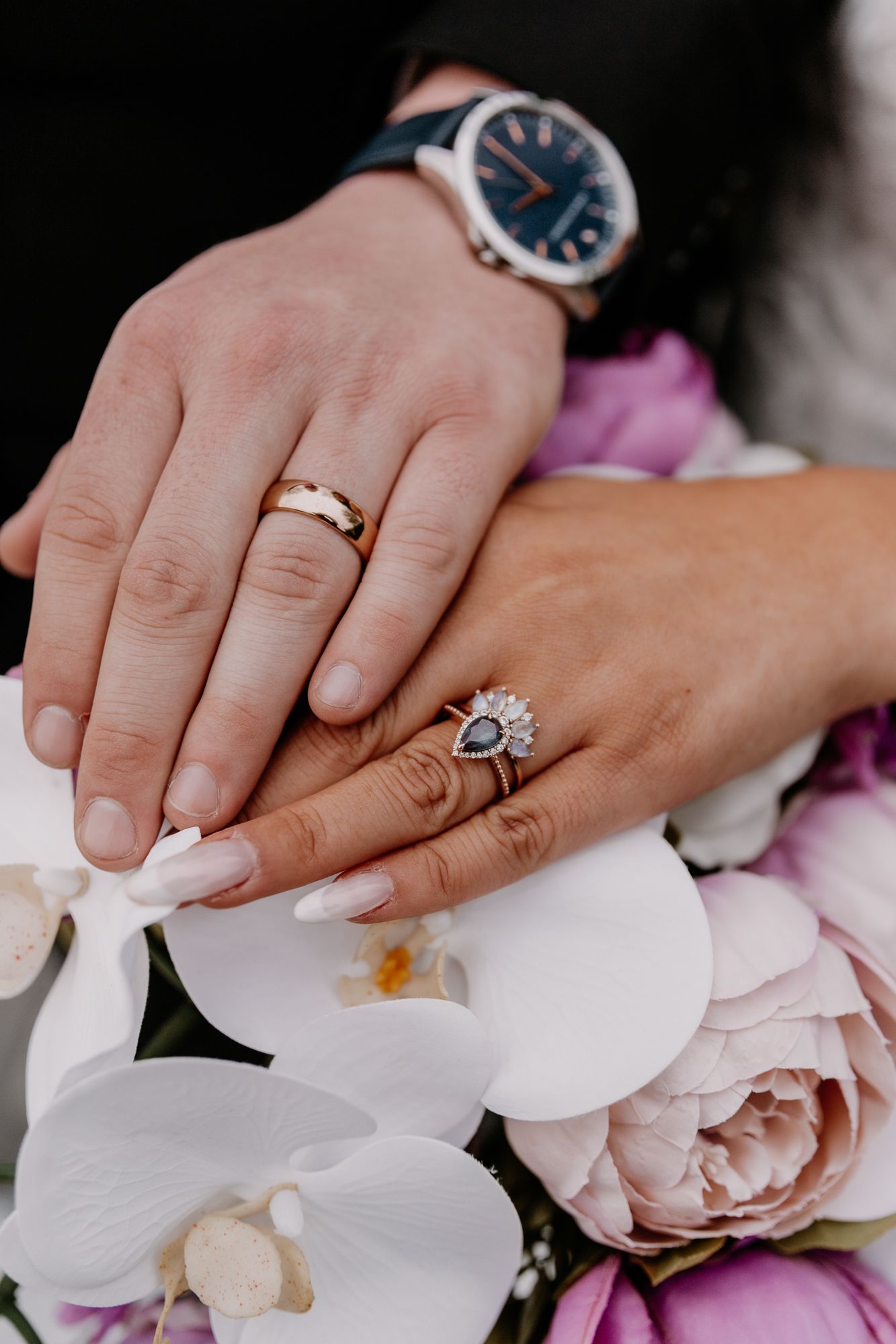 Wedding rings and watch detail shot with white orchids and pink roses in background.