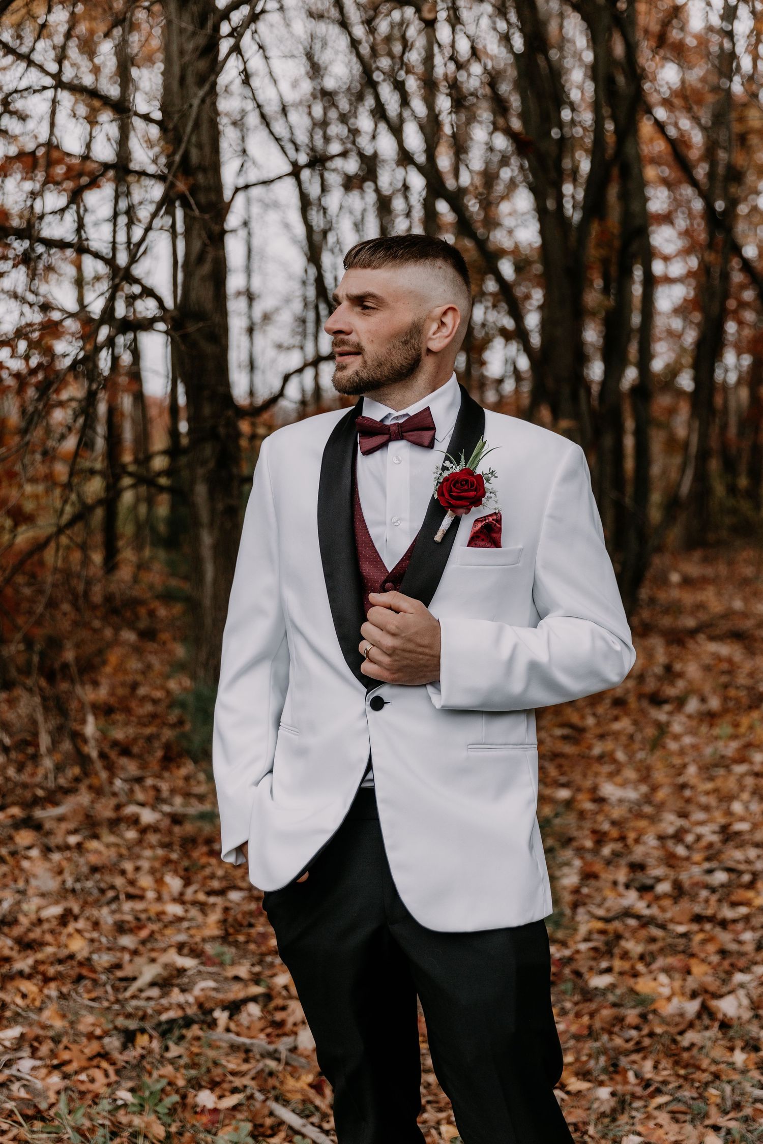 Groom in white tuxedo jacket and burgundy bow tie poses in autumn forest setting.